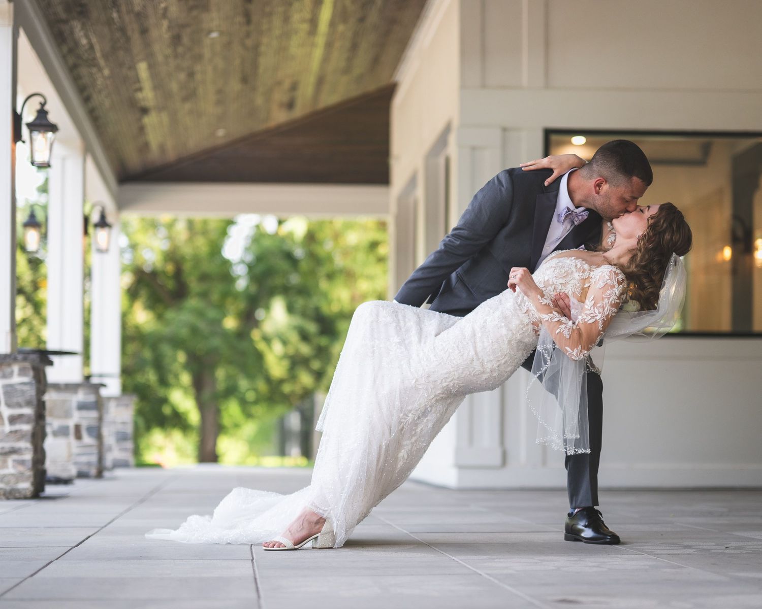 Groom in a dark suit dips and kisses his bride in a flowing white gown on a stone patio at their wedding venue.