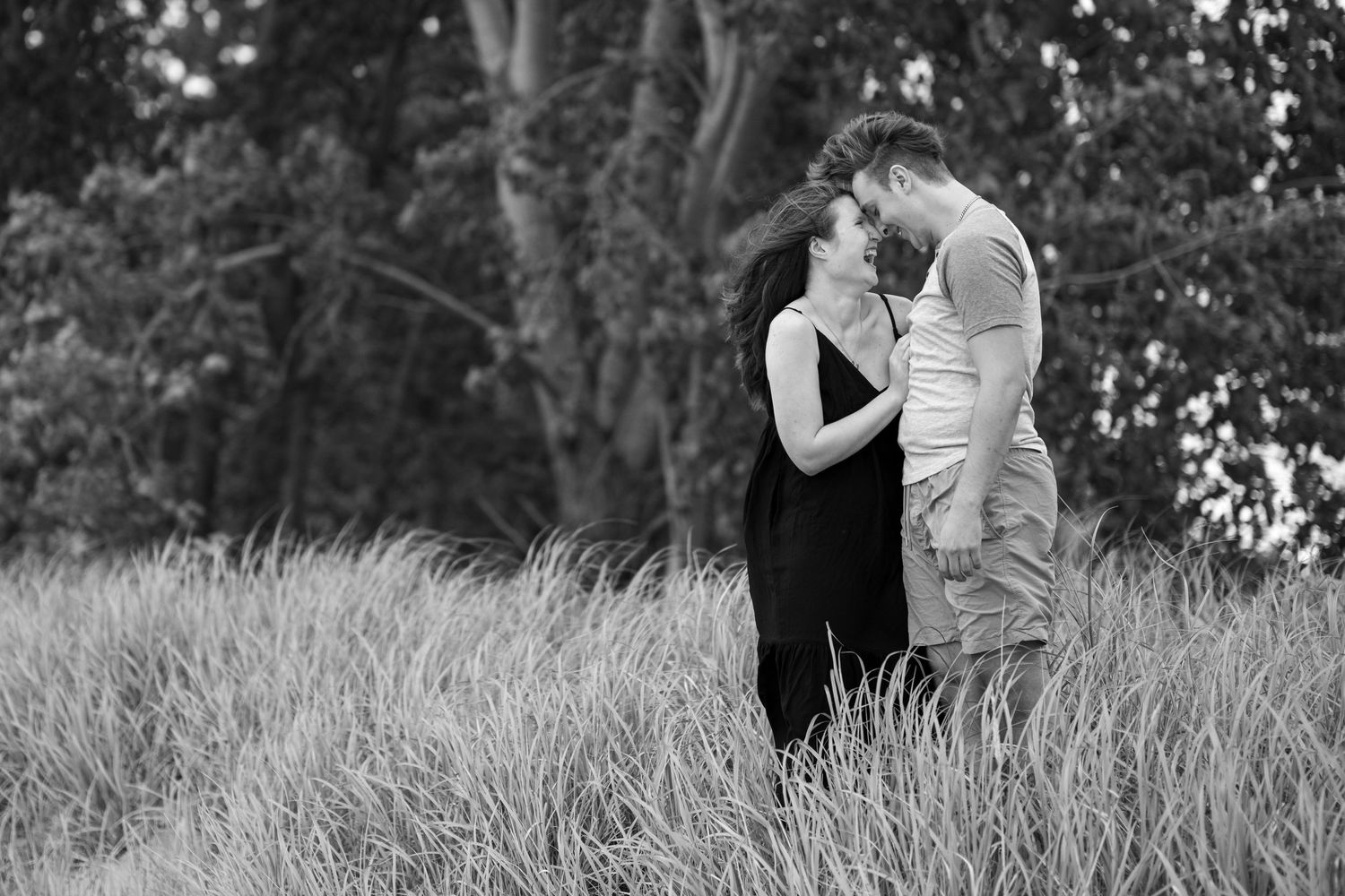 Black and white photo of a couple embracing and about to kiss in tall grass with trees in the background.