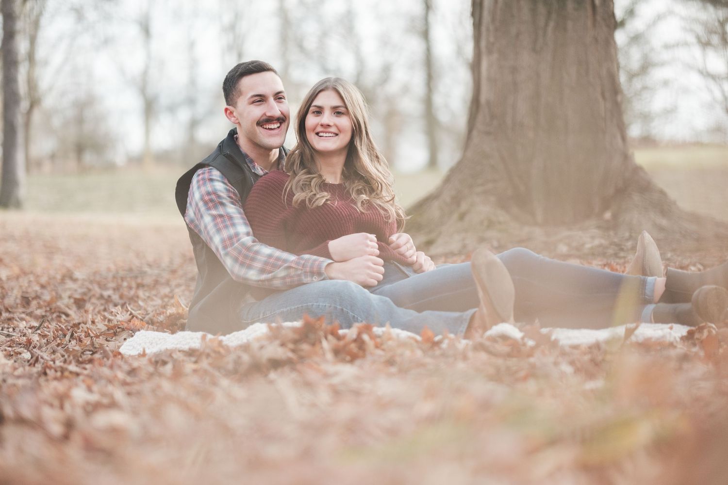 Smiling couple cuddling on a blanket among fallen autumn leaves beneath a large tree during an outdoor engagement photo session.