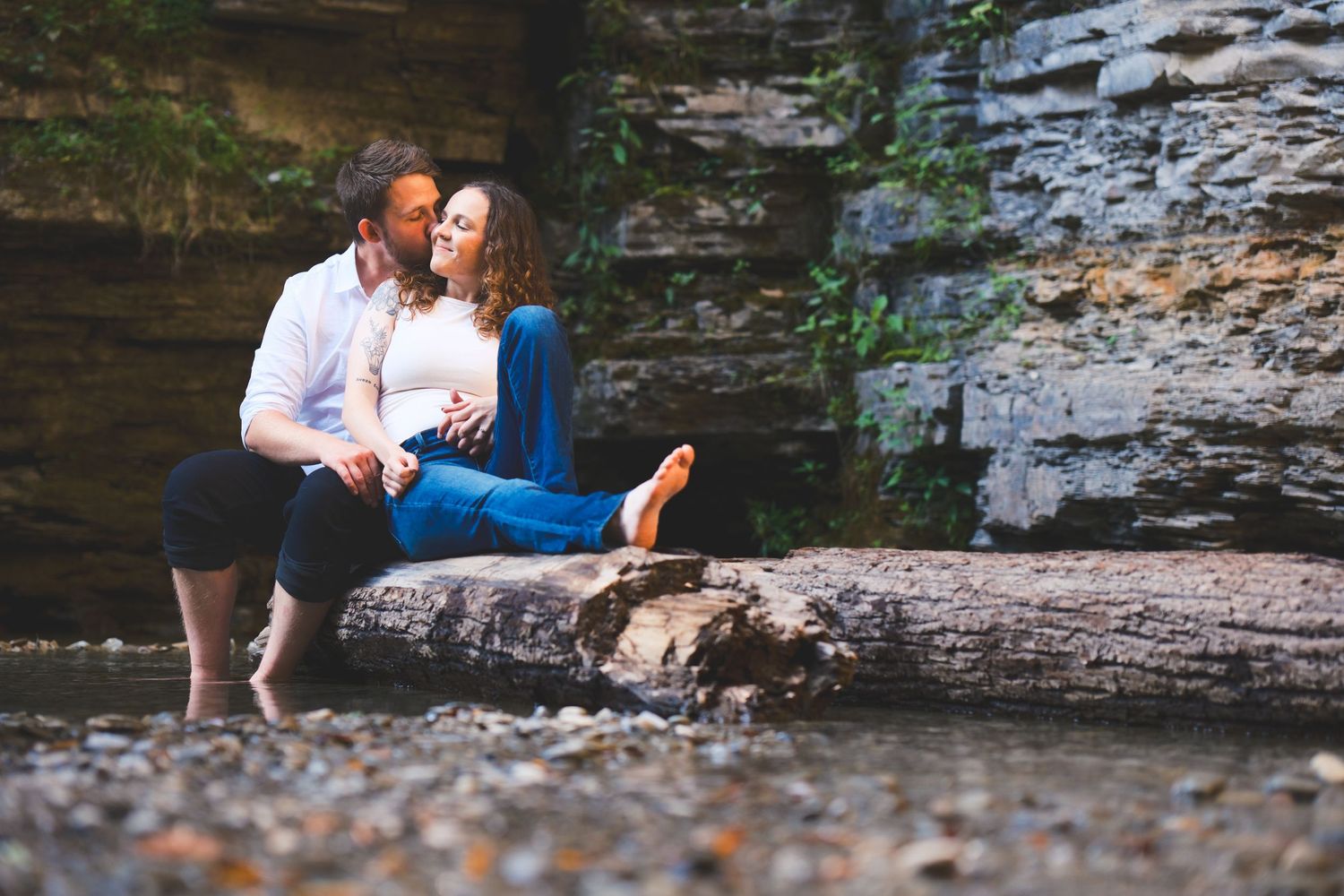 Couple sits on a fallen log beside a shallow creek and kisses in front of a rocky cliff during an outdoor engagement session.