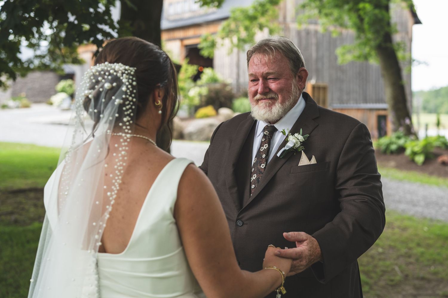 Smiling bearded groom in a brown suit and boutonniere holds the hand of his veiled bride in a white dress at an outdoor wedding.
