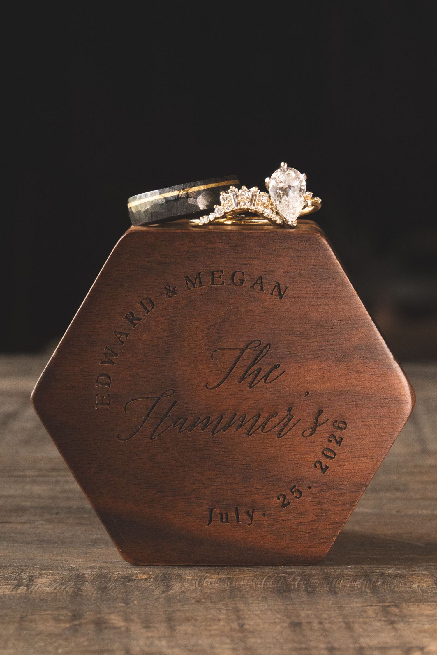 Engagement and wedding rings resting on an engraved hexagonal walnut ring box personalized with names and a July 25, 2026 wedding date.