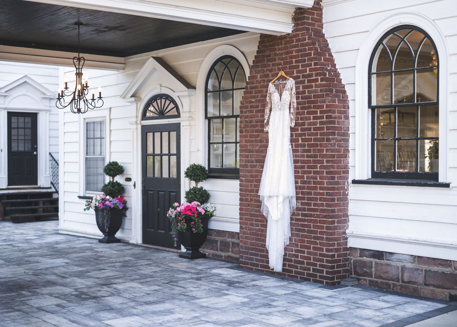 White wedding dress hangs from a hook on a brick column beside a white building with arched windows and flower planters.