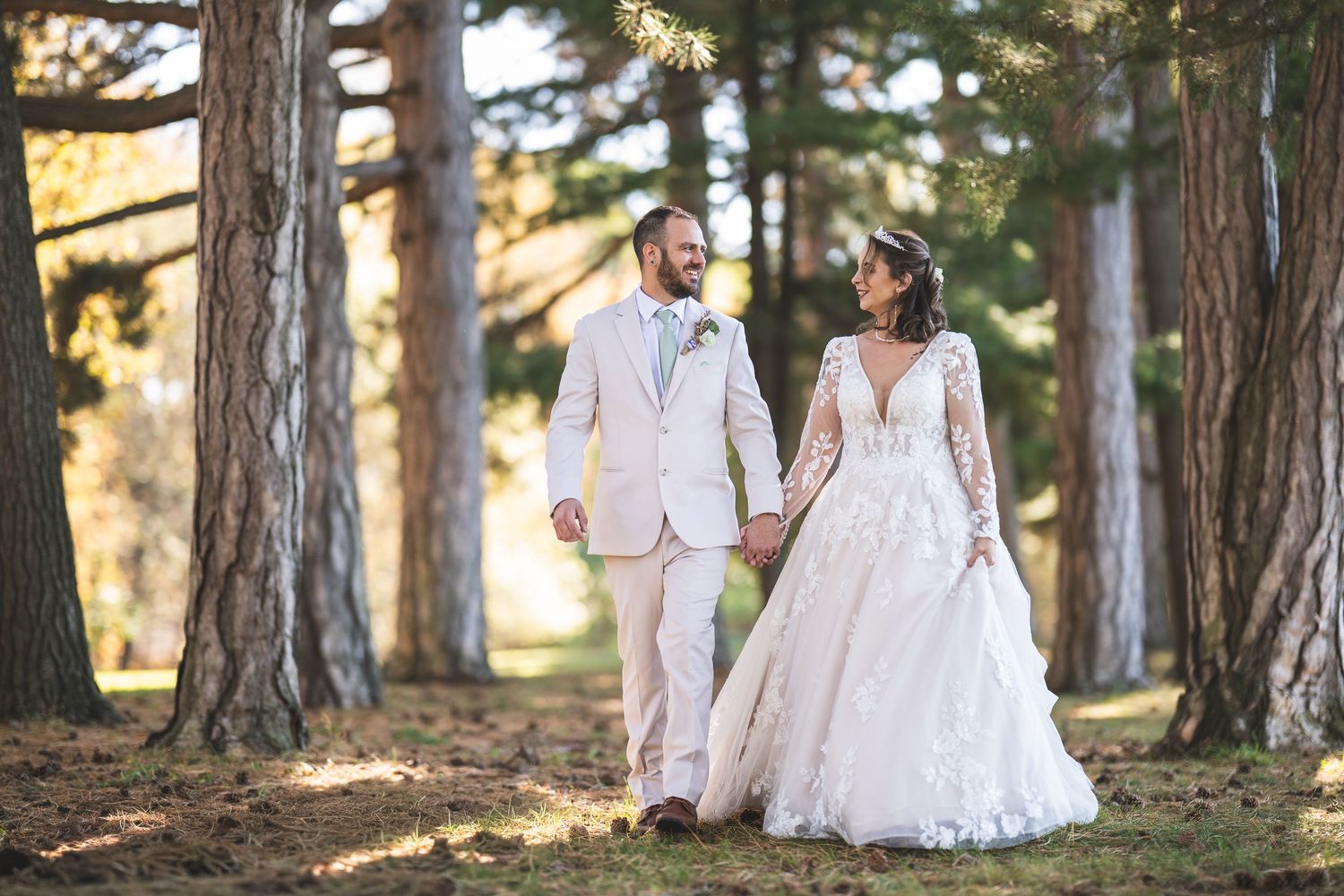 Bride in a lace wedding gown and groom in a light suit walk hand in hand through sunlit pine trees in a forest wedding portrait.
