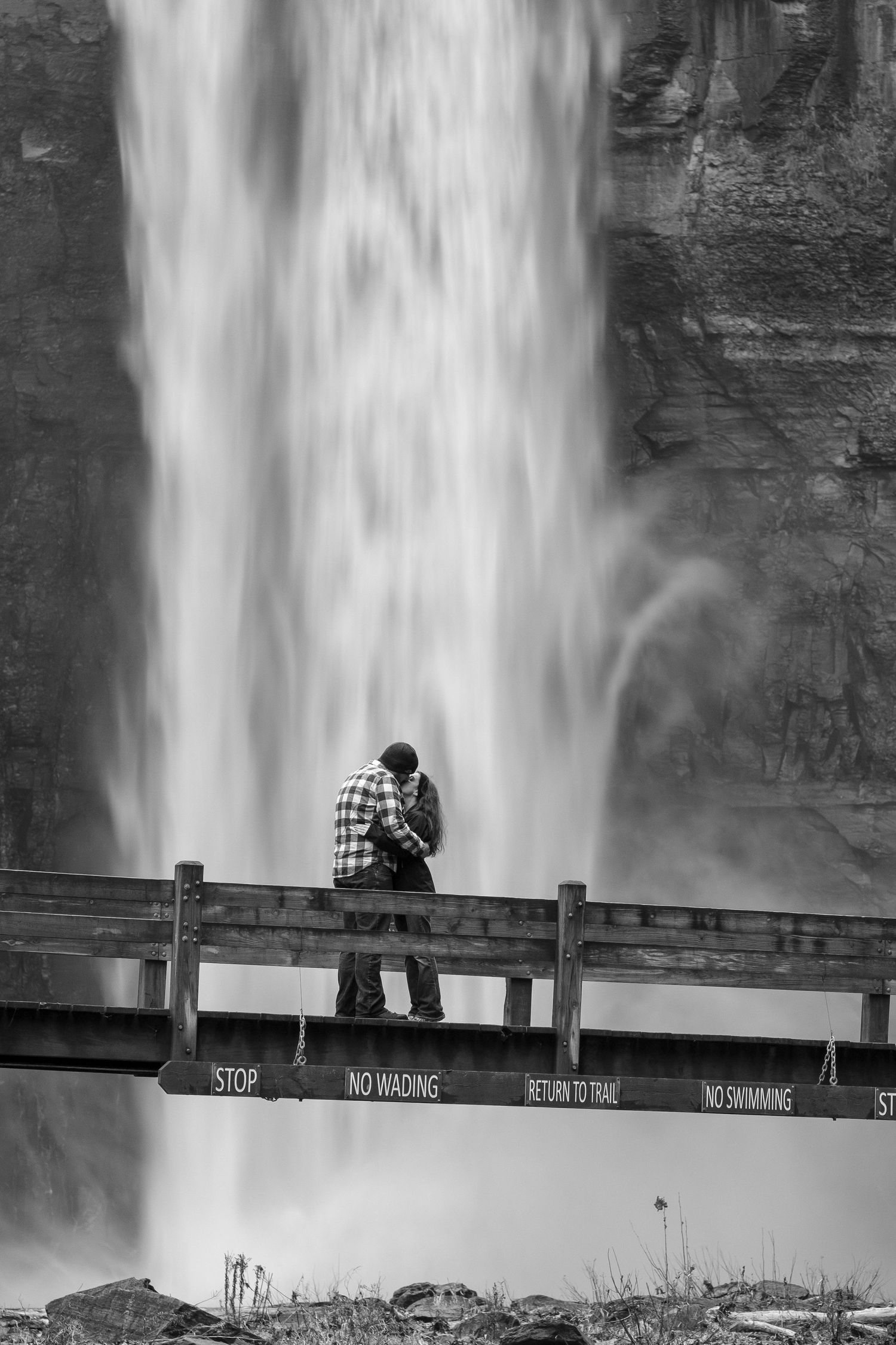 Monochrome image of a couple hugging on a wooden bridge beneath a tall waterfall, with warning signs along the railing.
