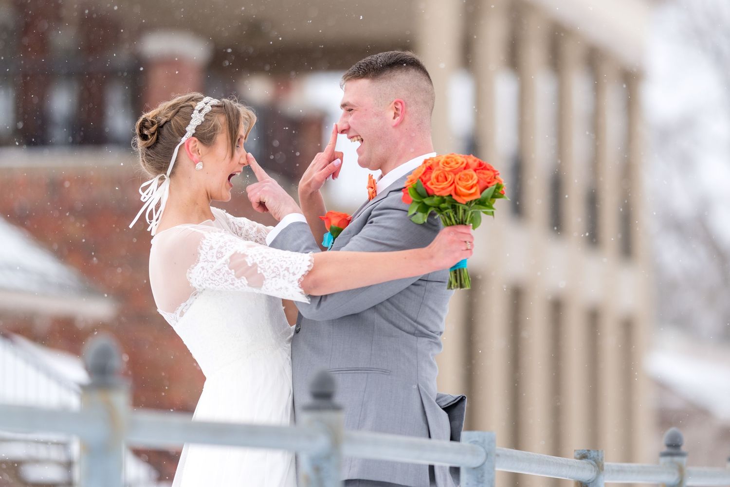 Smiling bride in a lace wedding dress and groom in a gray suit hold an orange bouquet and wipe happy tears during a snowy outdoor wedding.