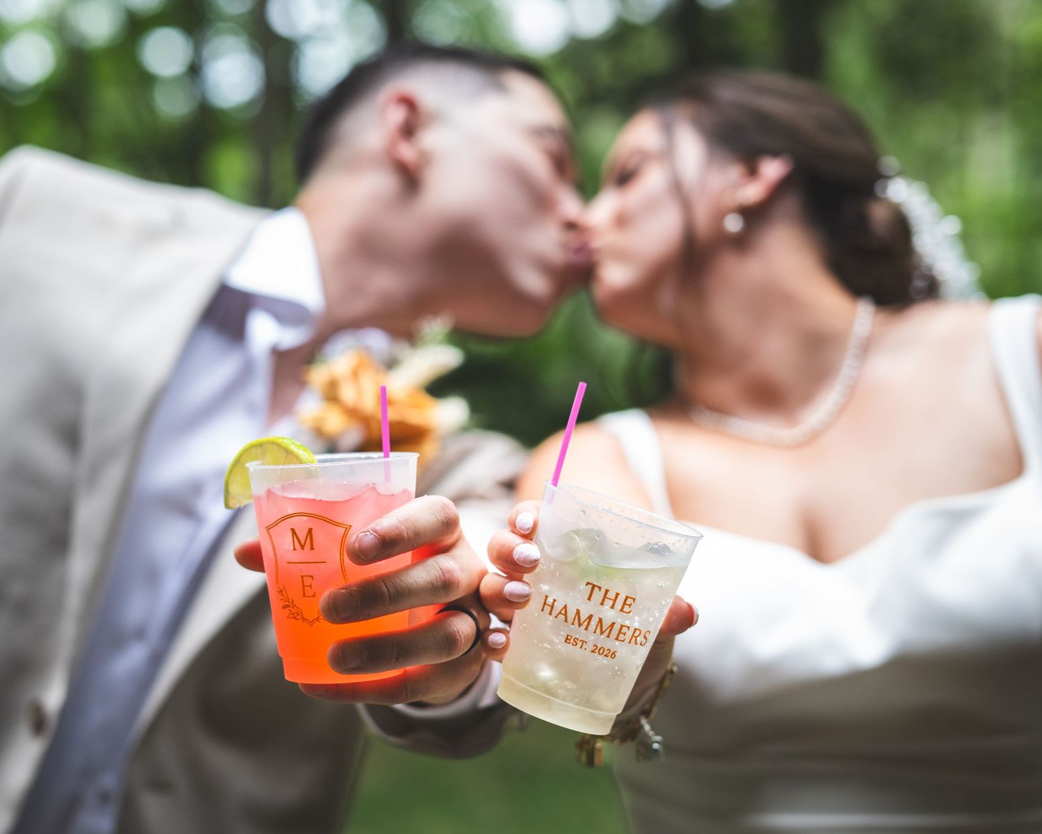 Bride and groom kiss outdoors while holding colorful cocktails in personalized cups with pink straws, surrounded by blurred green trees.