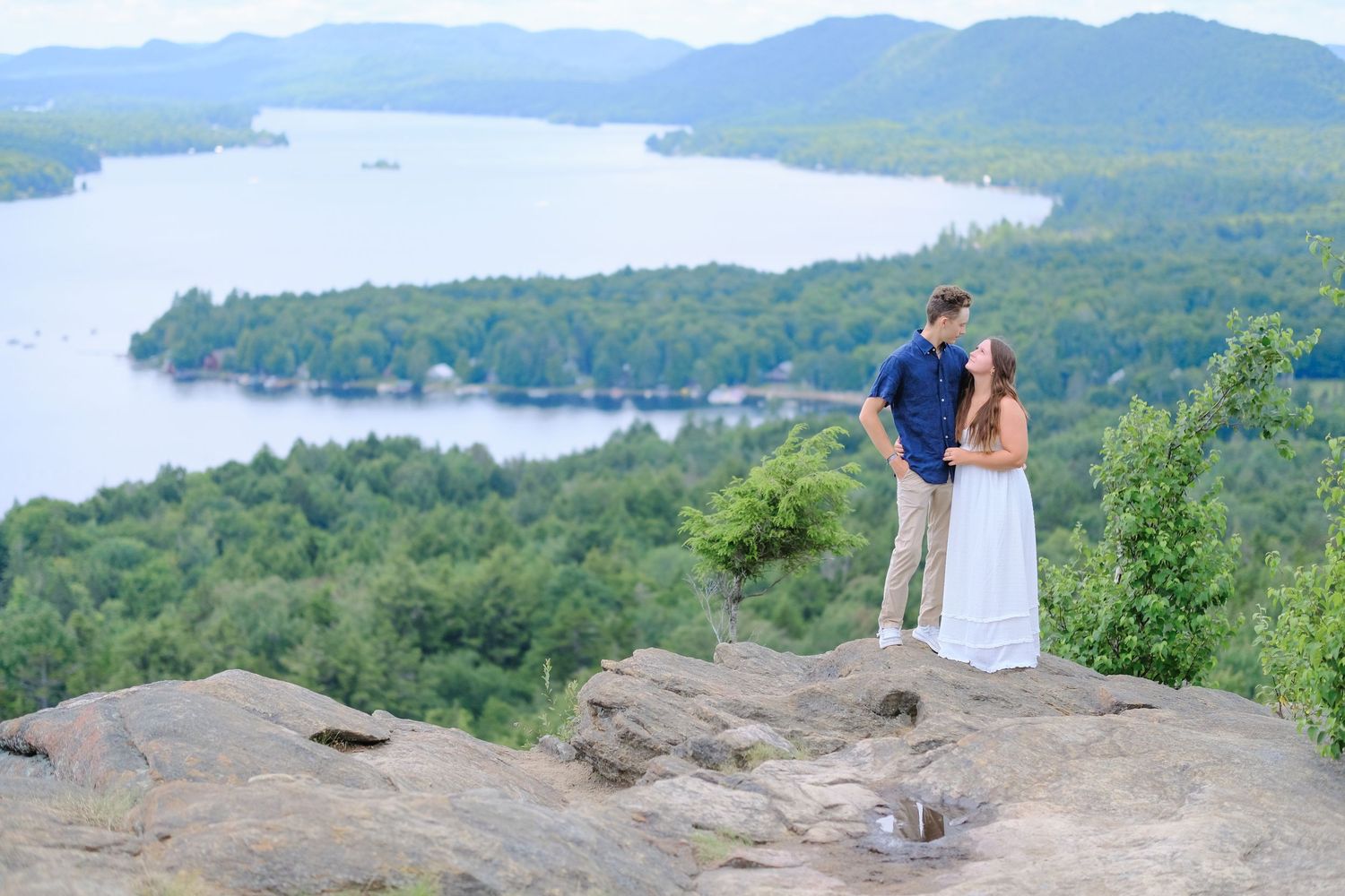 Couple embracing on a rocky mountain overlook with a scenic lake and forested hills stretching out behind them.