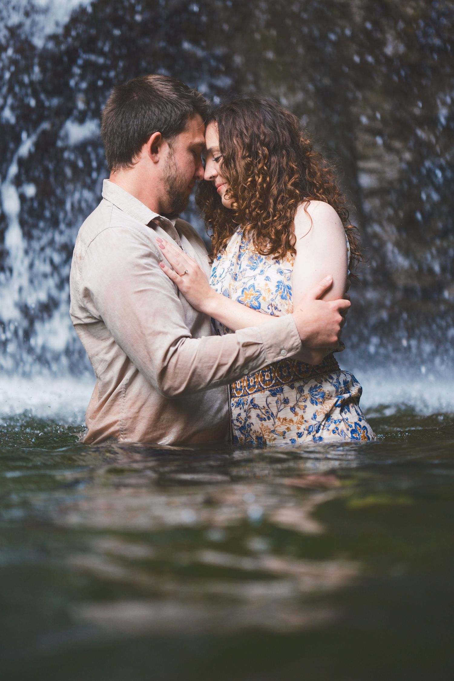 Romantic engagement photo of a couple embracing in a waterfall pool with misty spray and soft reflections on the water.