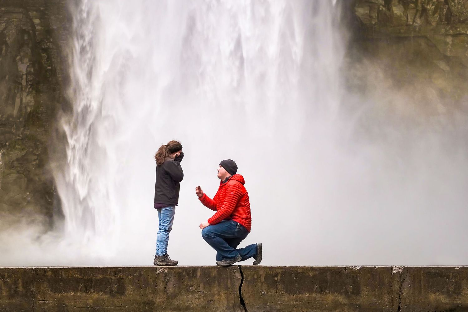 Man in a red jacket kneels to propose to his partner on a stone ledge in front of a towering, mist-shrouded waterfall.