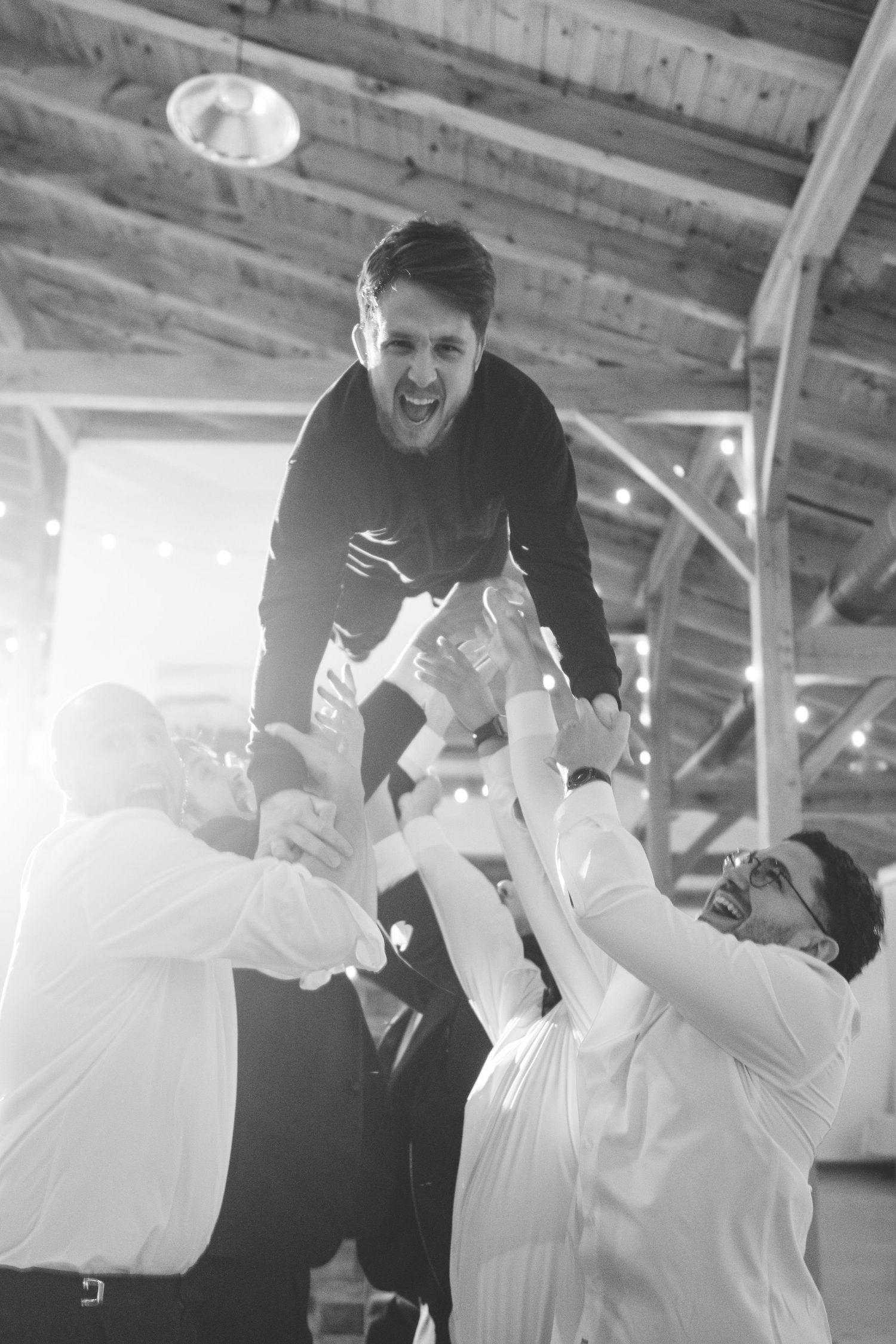 Wedding guests in white shirts lift a laughing man in the air during a lively celebration inside a rustic barn venue, black and white photo.