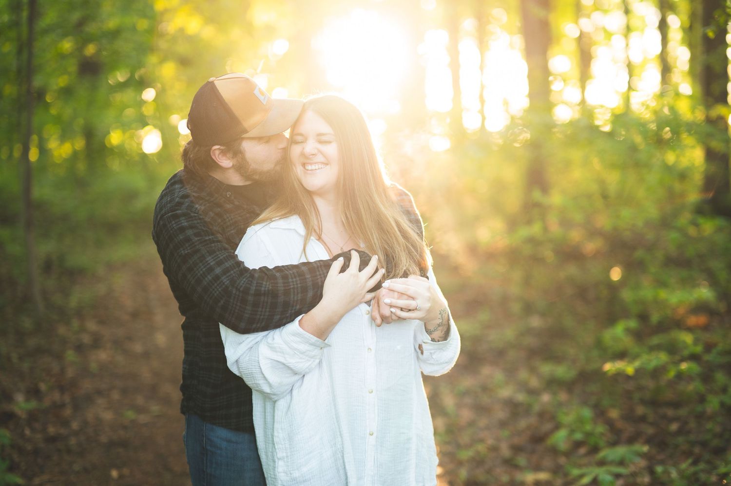 Smiling couple embracing in a sunlit forest at golden hour, the man hugging the woman in a white shirt from behind.