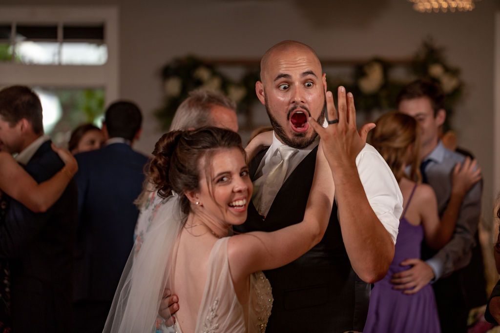 Joyful bride and groom dance at their wedding reception as the groom shows his ring with a playful open-mouthed expression.