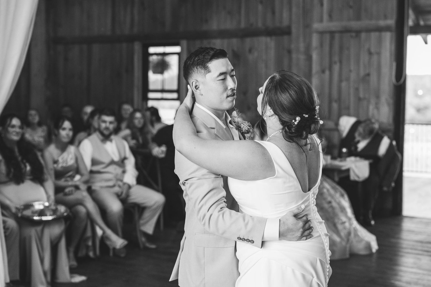 A black and white photo of a bride and groom sharing a first dance in a rustic barn as wedding guests look on.