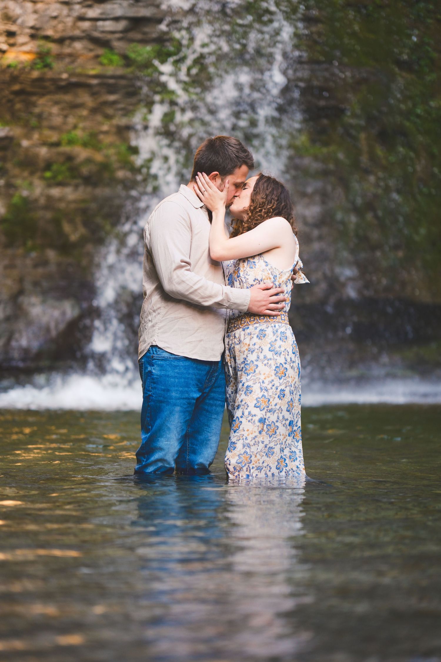 Couple in a floral dress and button-up shirt kiss while standing waist-deep in water in front of a waterfall.