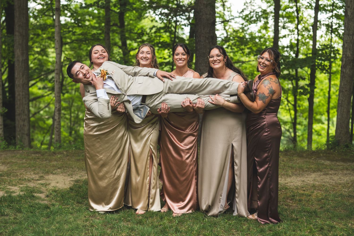 Five bridesmaids in satin gowns laugh while holding a groom in a gray suit horizontally in their arms on a grassy forest clearing.