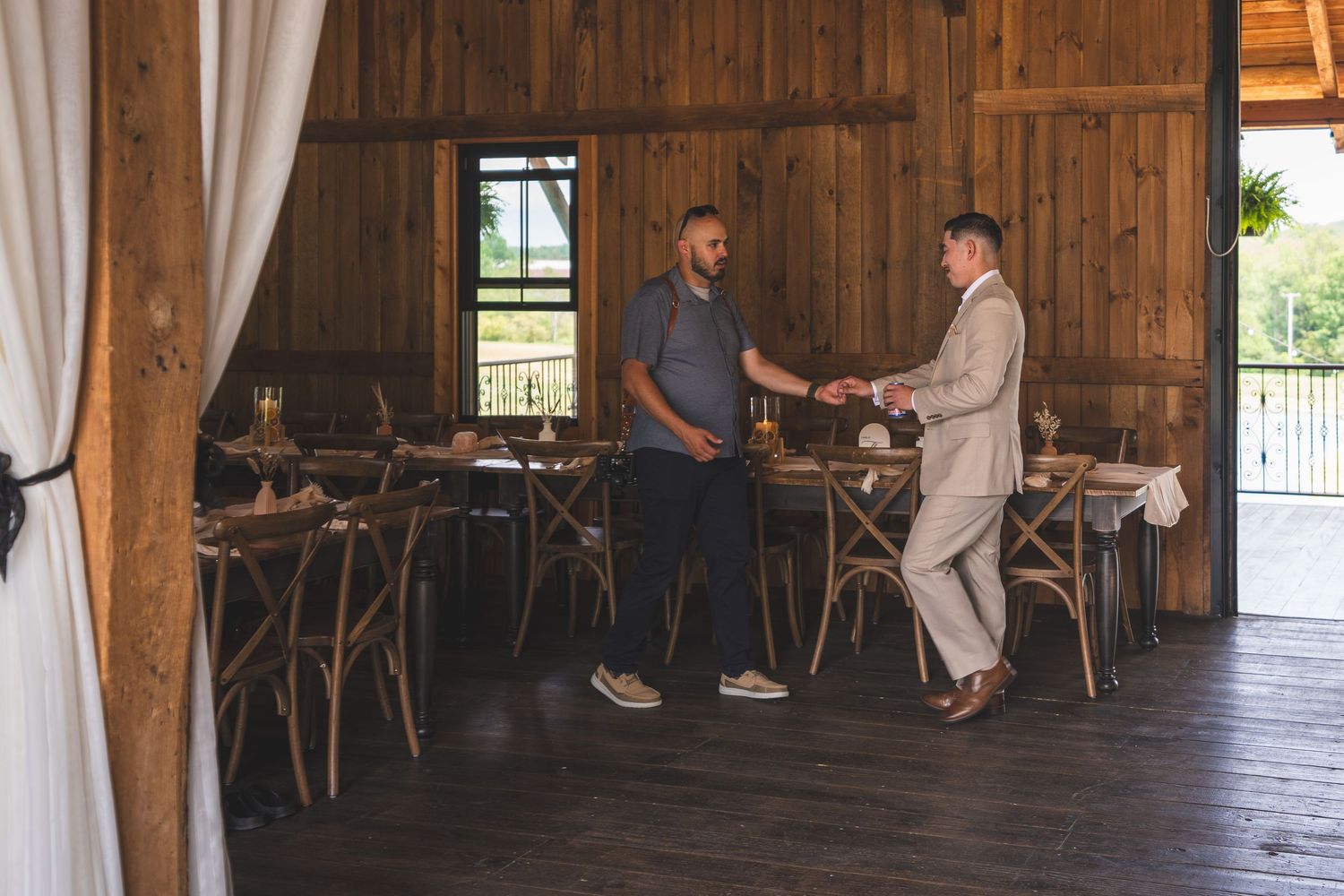 Photographer shakes hands with a man in a light suit inside a rustic wooden barn venue set with tables and chairs for a wedding.