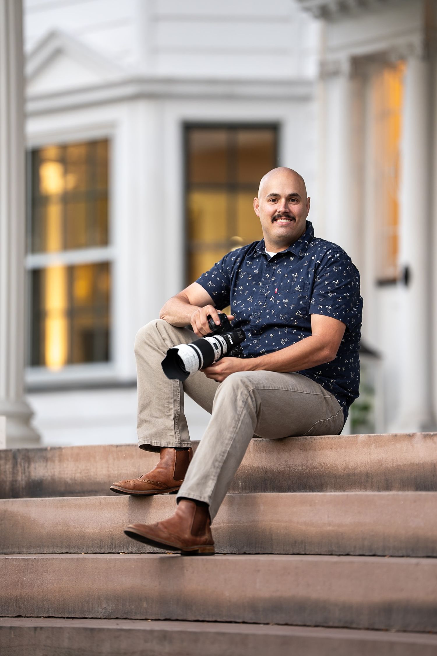 Smiling man in a navy floral shirt and khaki pants sits on stone steps holding a camera with a telephoto lens outside a building.