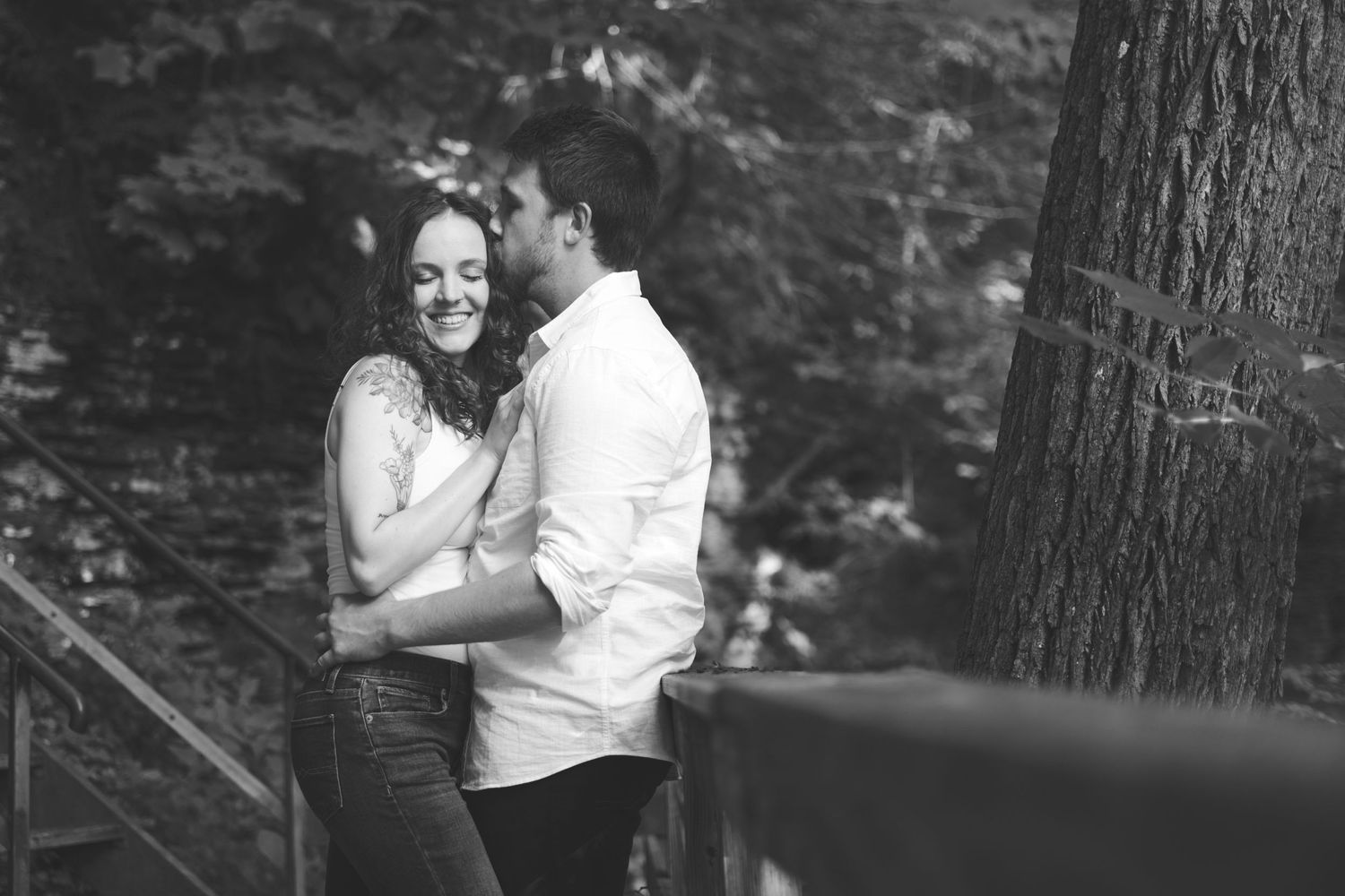 Black and white engagement photo of a couple embracing and smiling beside a large tree and wooden railing in a wooded park.