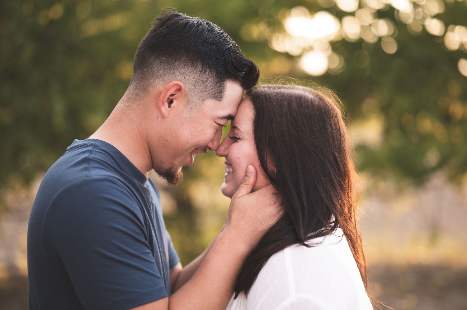 A smiling couple stands forehead to forehead outdoors, the woman gently touching the man's face, with a softly blurred green background.