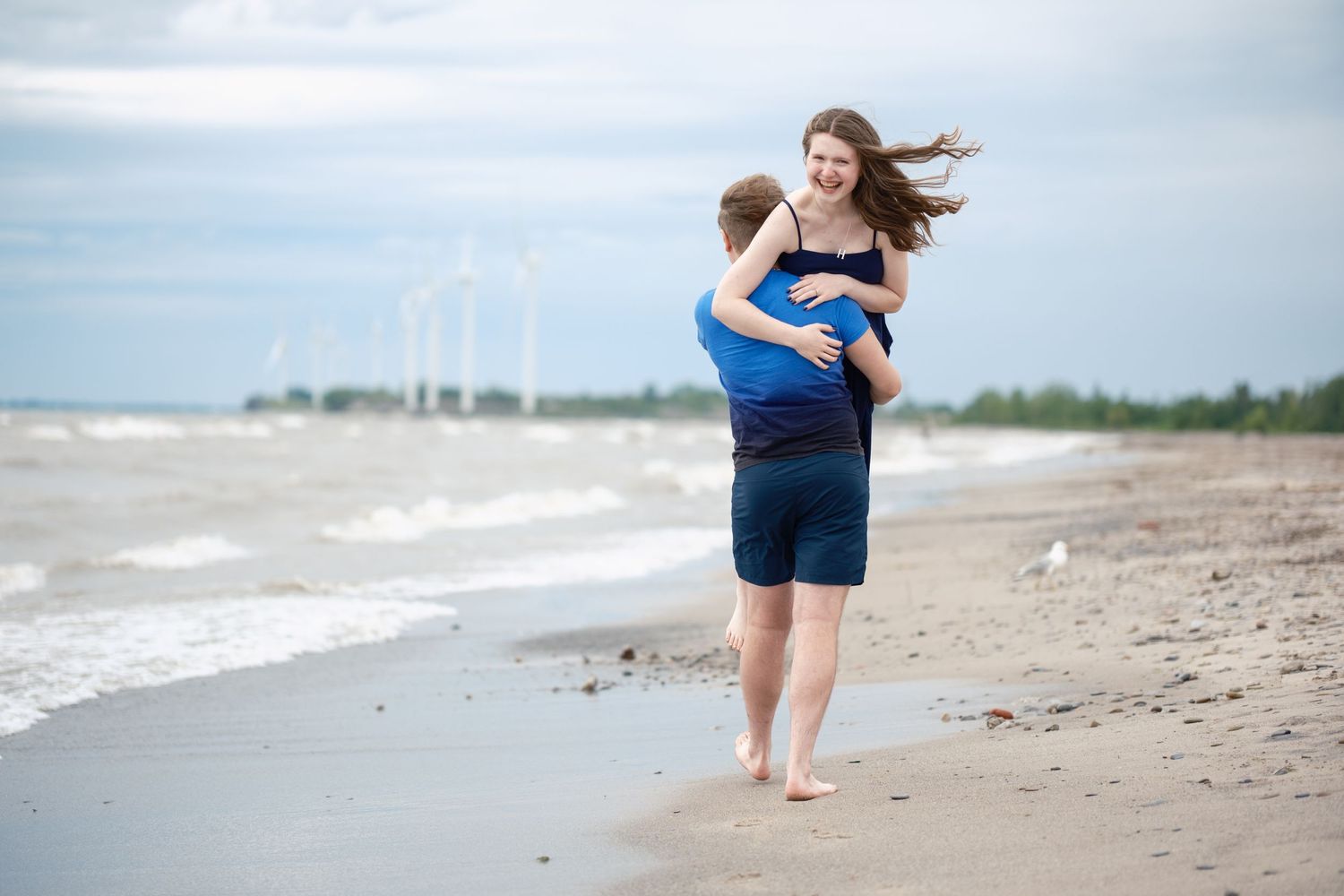 Two people embrace while standing barefoot on a windy beach with waves and windmills visible in the distance.