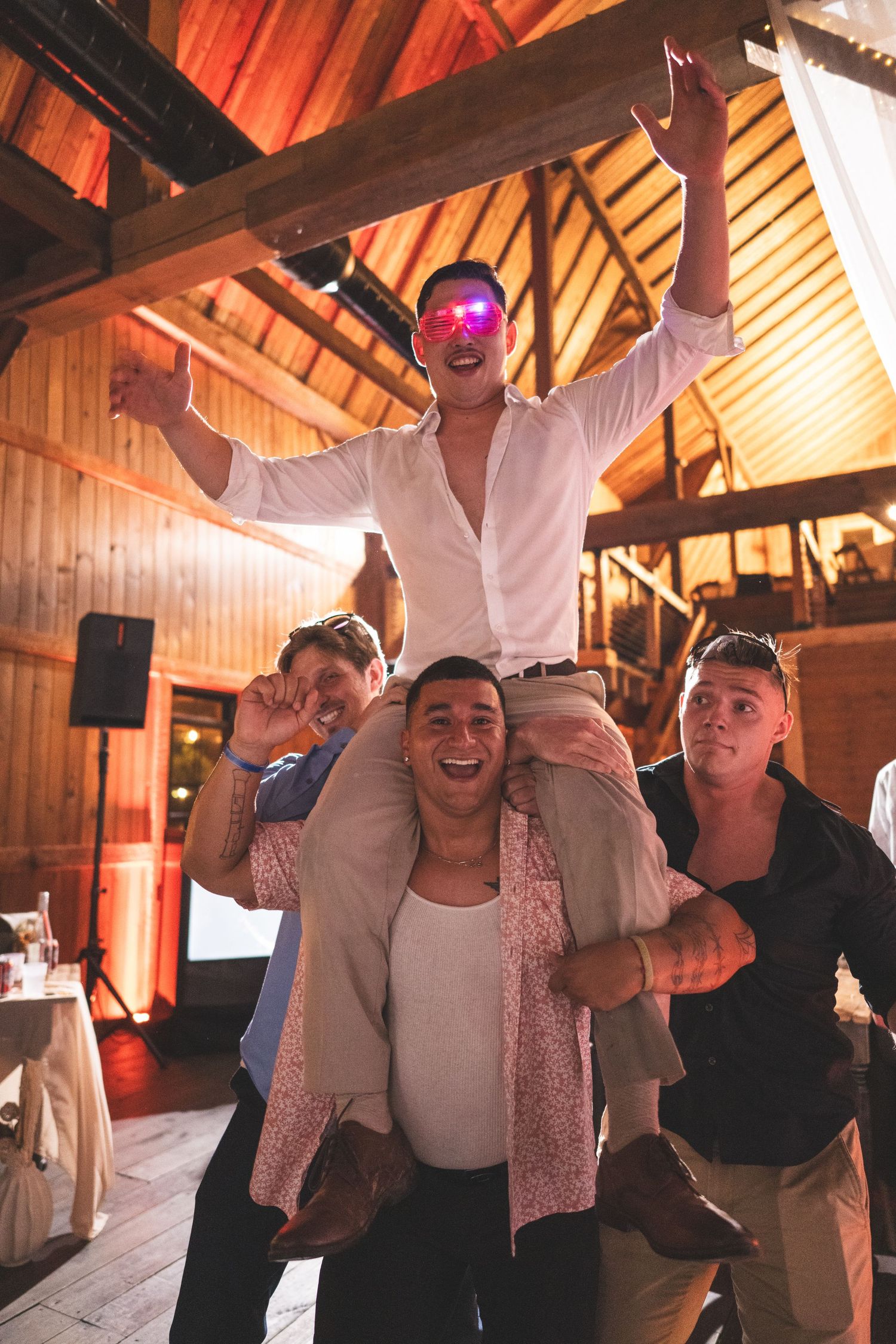 Man in white shirt and pink sunglasses cheers on a friend's shoulders at a lively party inside a rustic wooden barn venue.