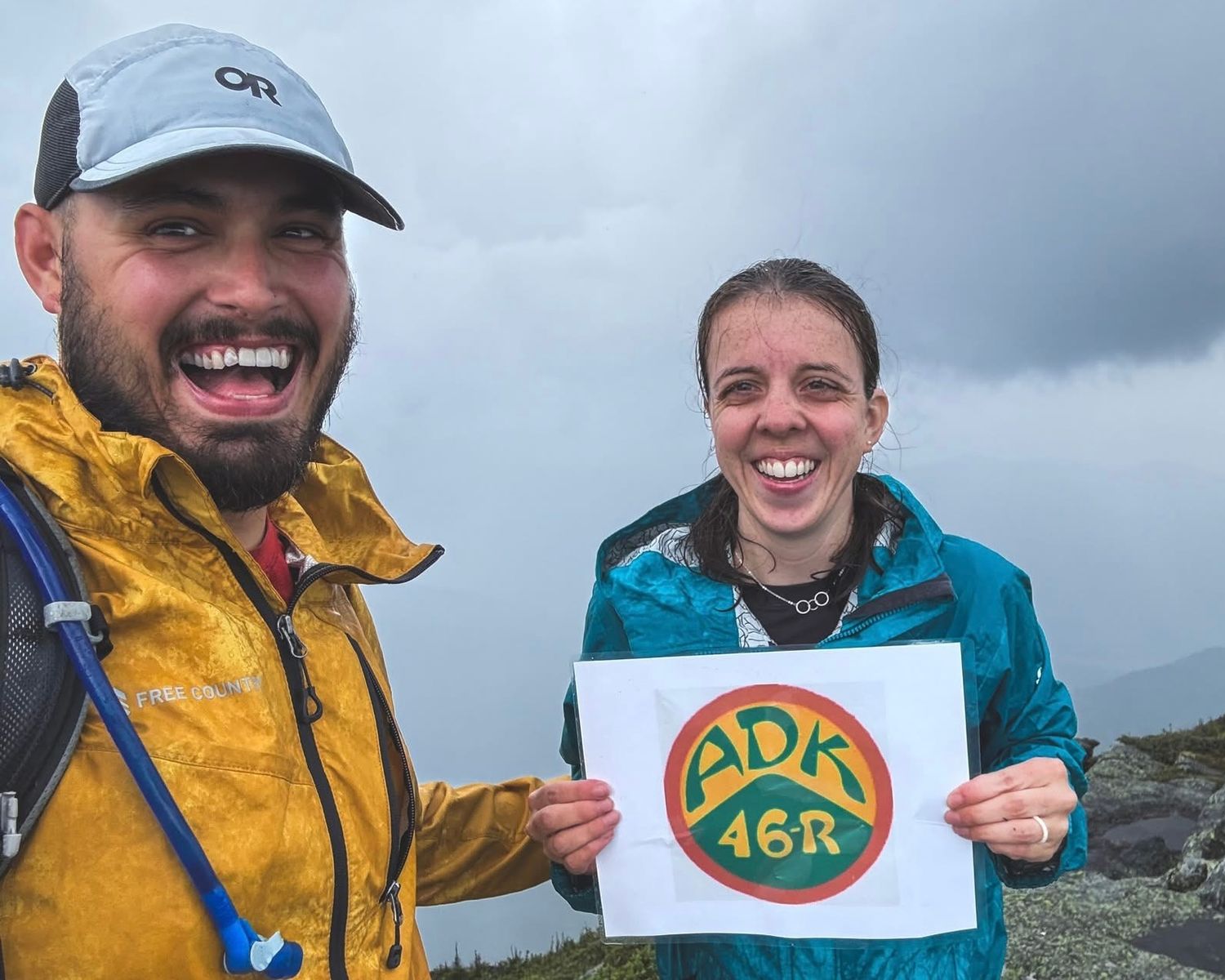 Two hikers in rain jackets laugh while holding an ADK 46R sign at a misty Adirondack summit after completing the 46 High Peaks.