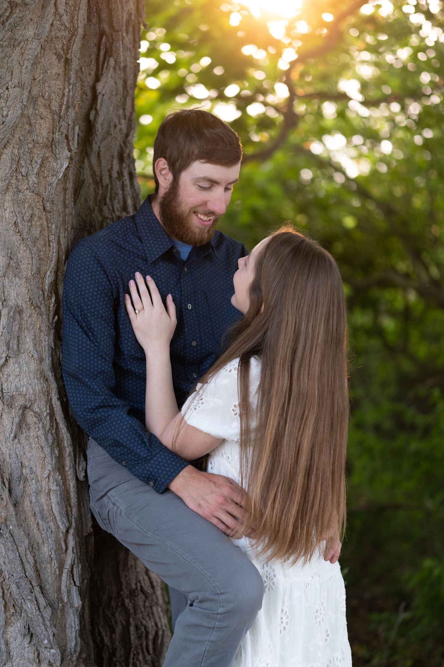 A bearded man in a navy shirt leans against a large tree trunk as a long-haired woman in a white dress embraces him in golden sunset light.