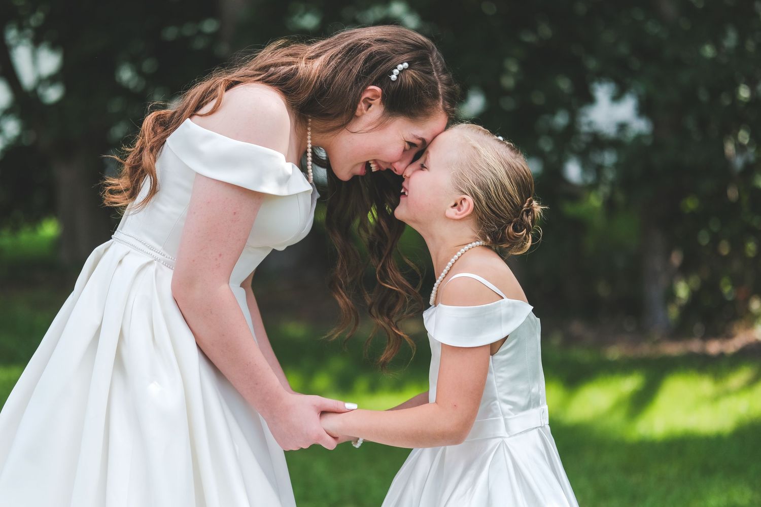 A bride in a white off-the-shoulder gown and a young flower girl in a matching dress touch foreheads and smile while holding hands outdoors.