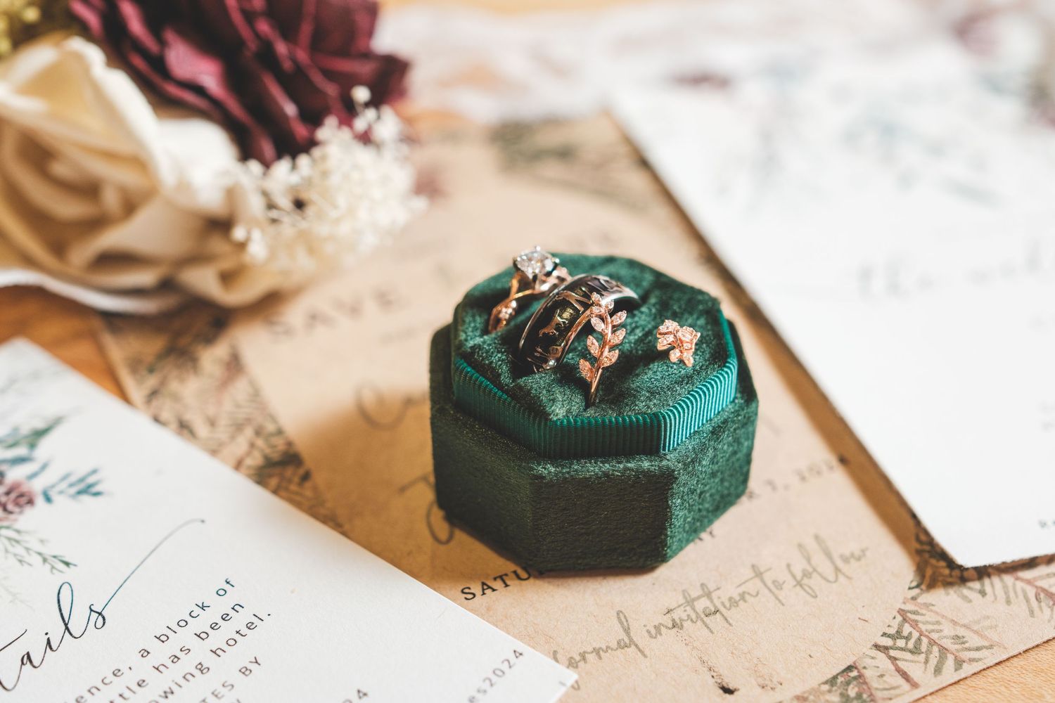 Open green velvet ring box holding ornate gold wedding bands on rustic wedding invitations beside burgundy and cream flowers.