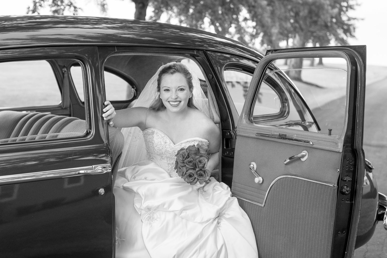 Smiling bride in a strapless wedding gown holding a bouquet while seated in a vintage car with the door open, black and white photo.