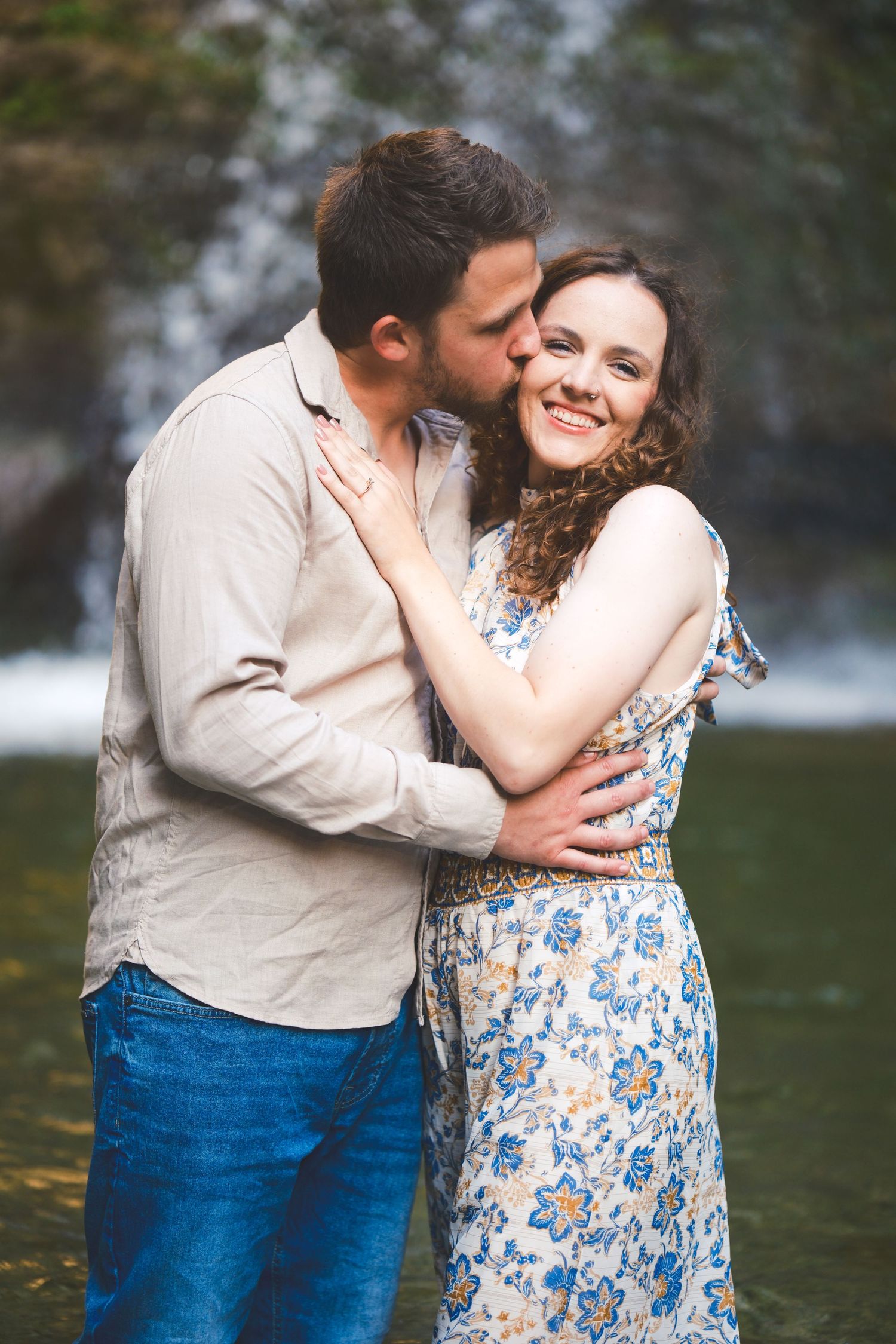 Smiling woman in a floral dress is kissed on the cheek by her fiance while they stand in water near a waterfall.