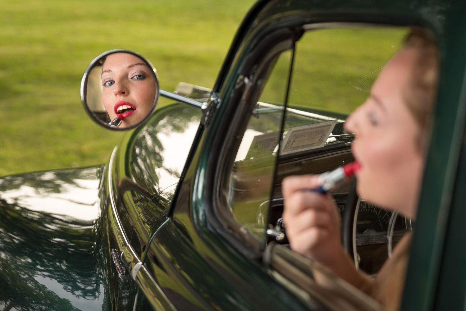 Woman applying red lipstick while looking into the side mirror of a vintage green car parked on a grassy field.