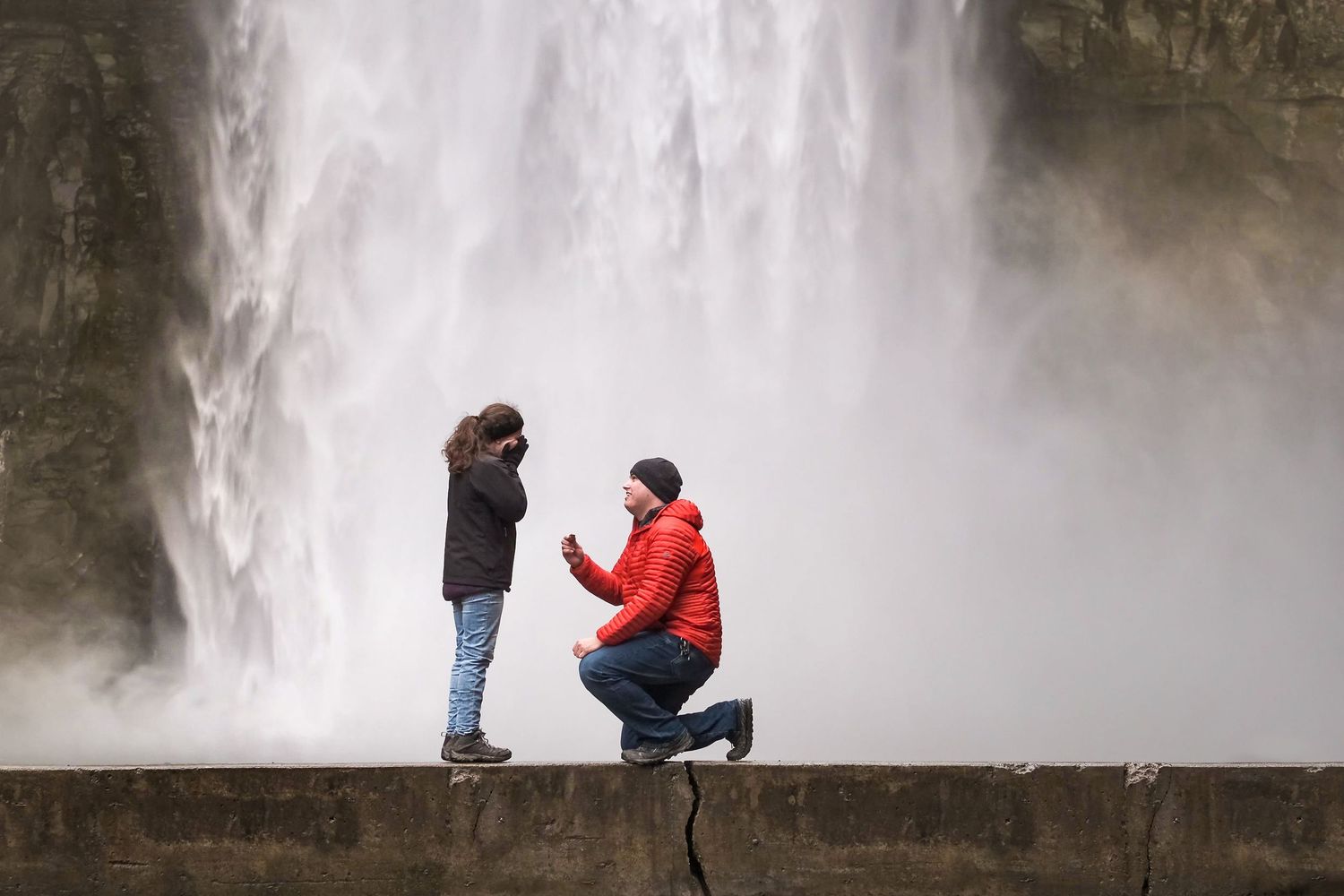 Man in a red jacket kneels on one knee proposing to a woman on a stone ledge in front of a large, misty waterfall.