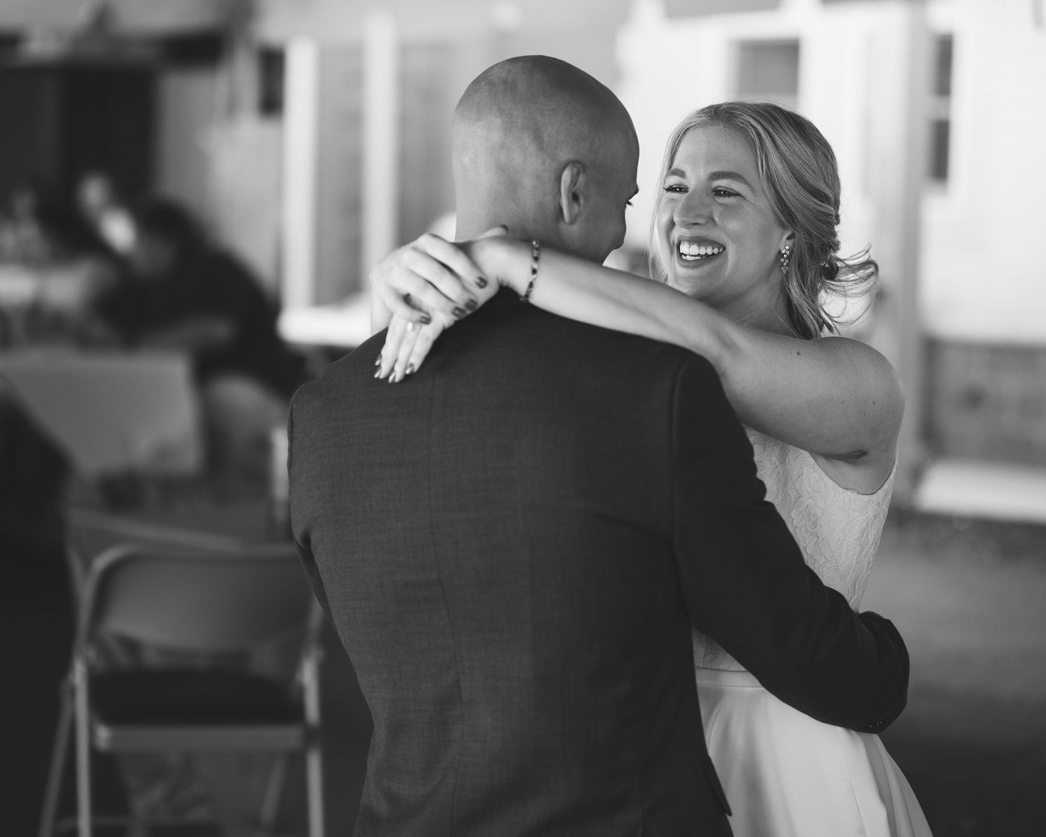 Smiling bride in a white dress embraces a bald groom in a dark suit during a joyful wedding first dance, in black and white.