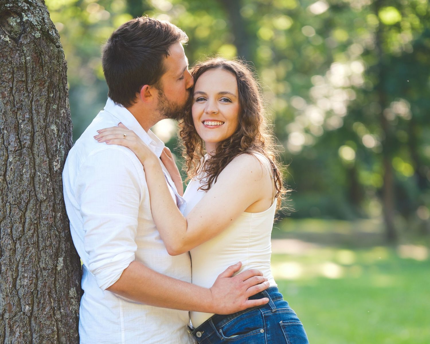 A man kisses a smiling woman on the cheek as they lean against a tree in a sunlit park for an engagement portrait.