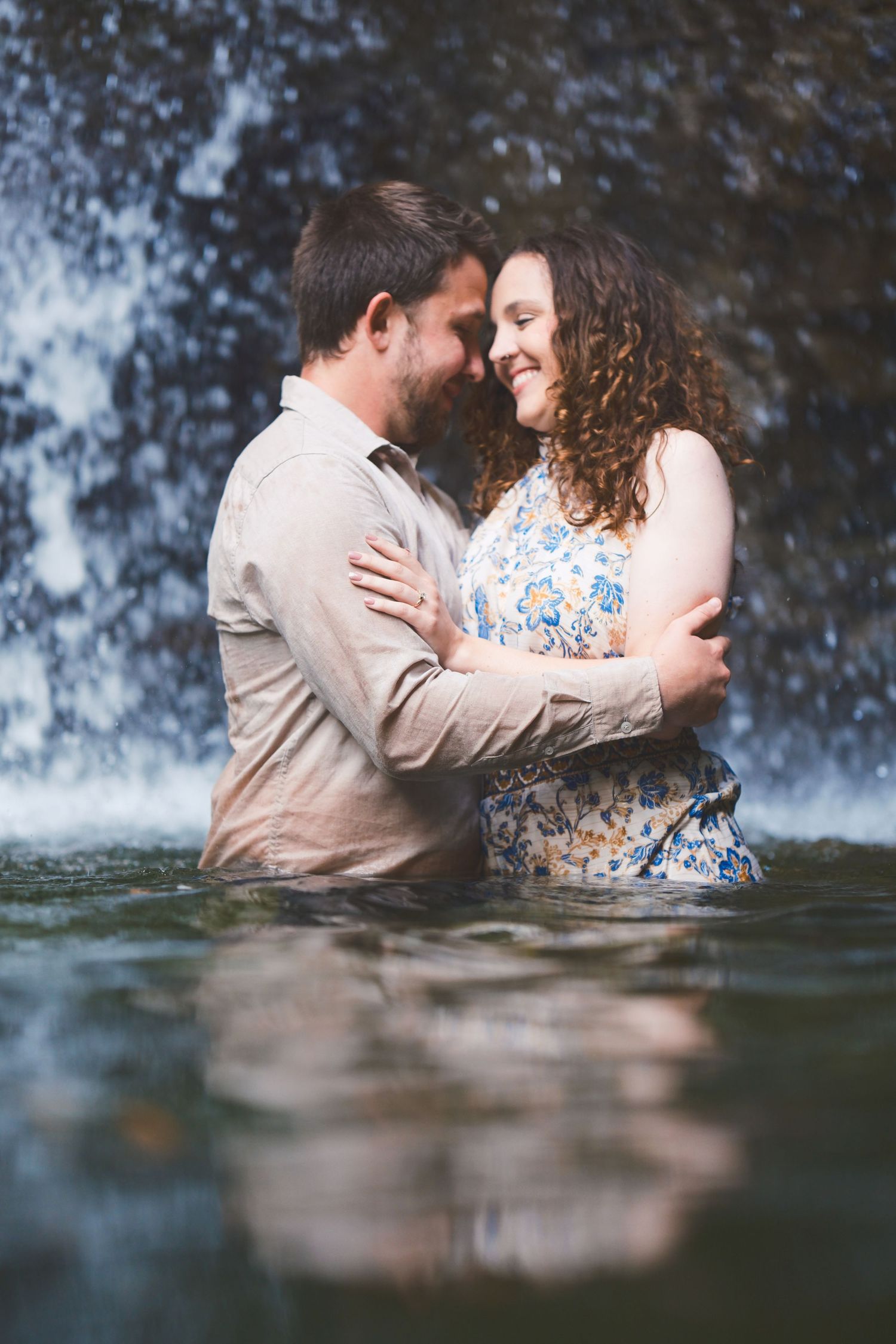 Couple holds each other close and smile while standing in water with waterfall spray behind them in an engagement photo.
