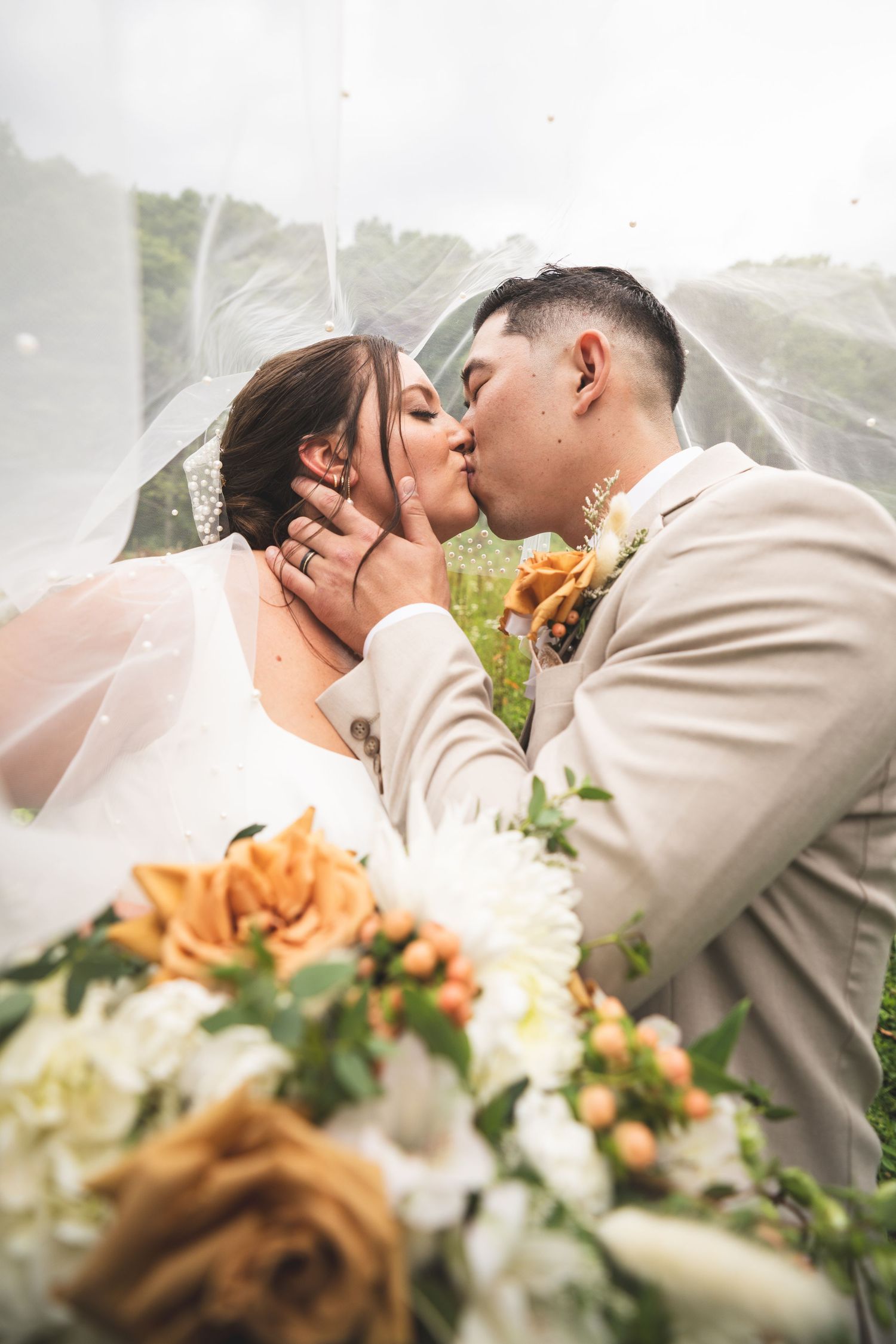 Bride and groom share a kiss under a sheer veil, framed by orange and white wedding bouquet flowers with a green hillside behind.