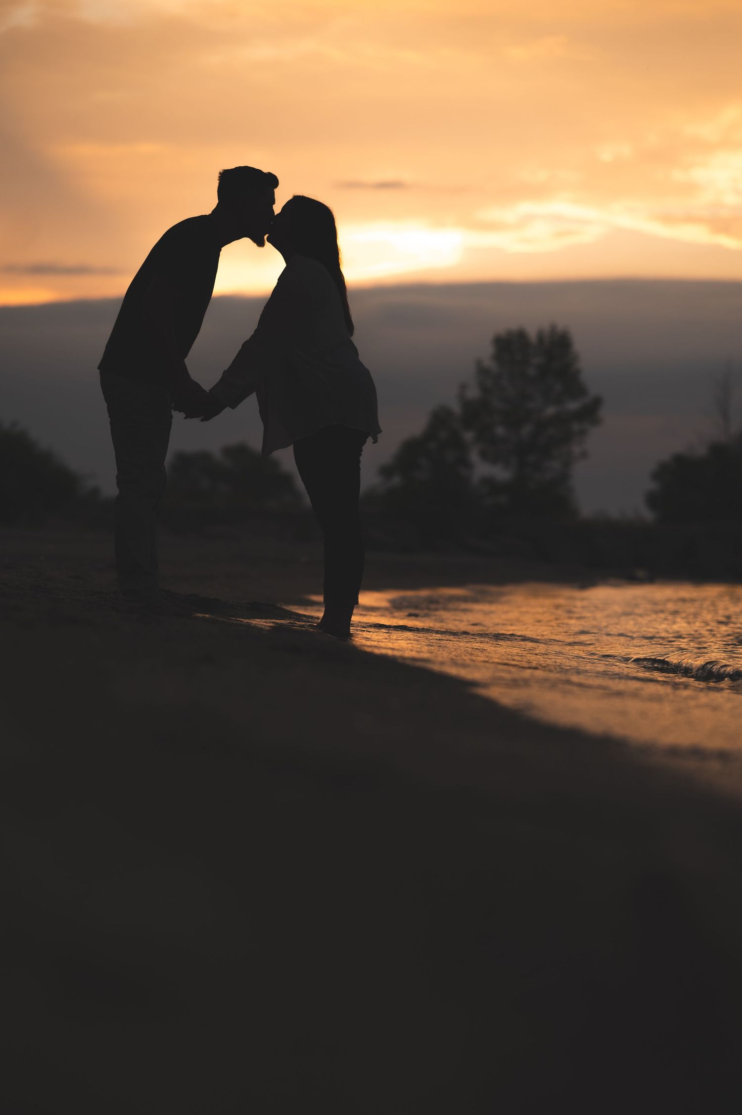 Silhouette of a couple kissing while holding hands on a sandy beach at sunset, with orange sky and calm water behind them.