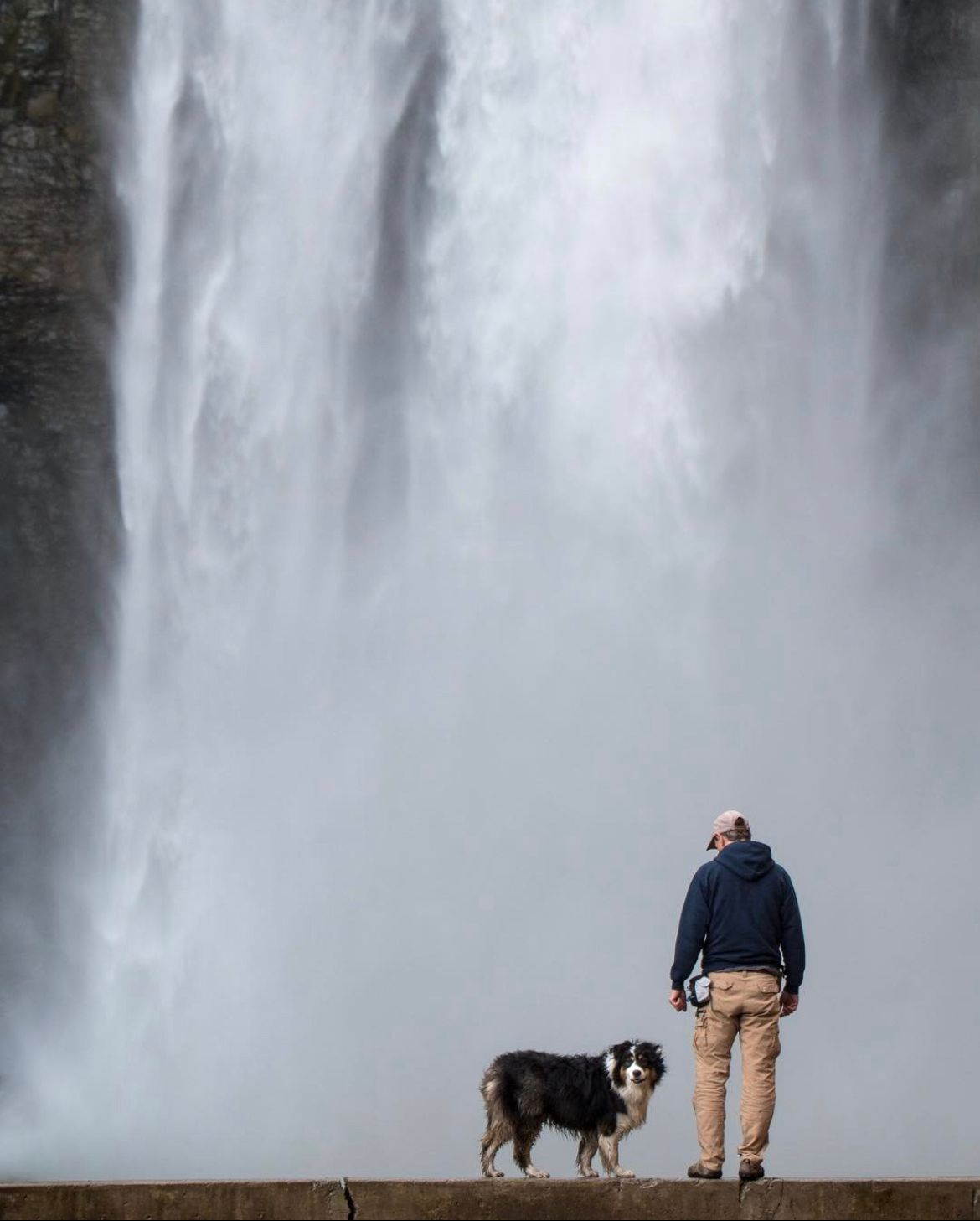 Man in a navy hoodie stands with an Australian shepherd dog on a stone ledge, facing a huge misty waterfall.