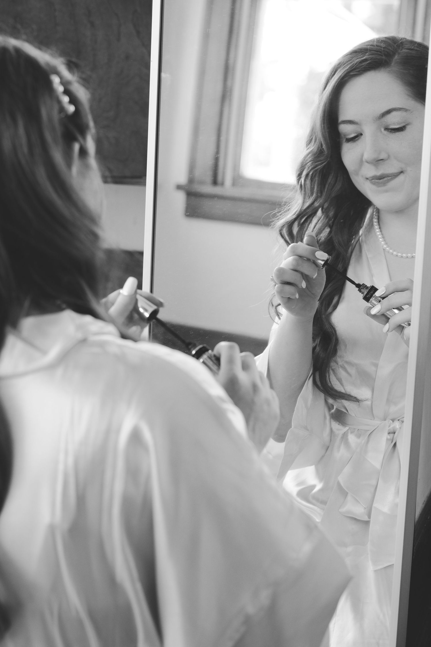 Black and white photo of a woman in a satin robe and pearl necklace applying mascara in a mirror while getting ready.
