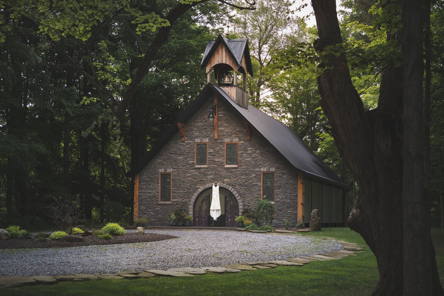 Rustic stone chapel with a wooden bell tower and arched entrance, surrounded by tall trees and a gravel path in a peaceful forest setting.