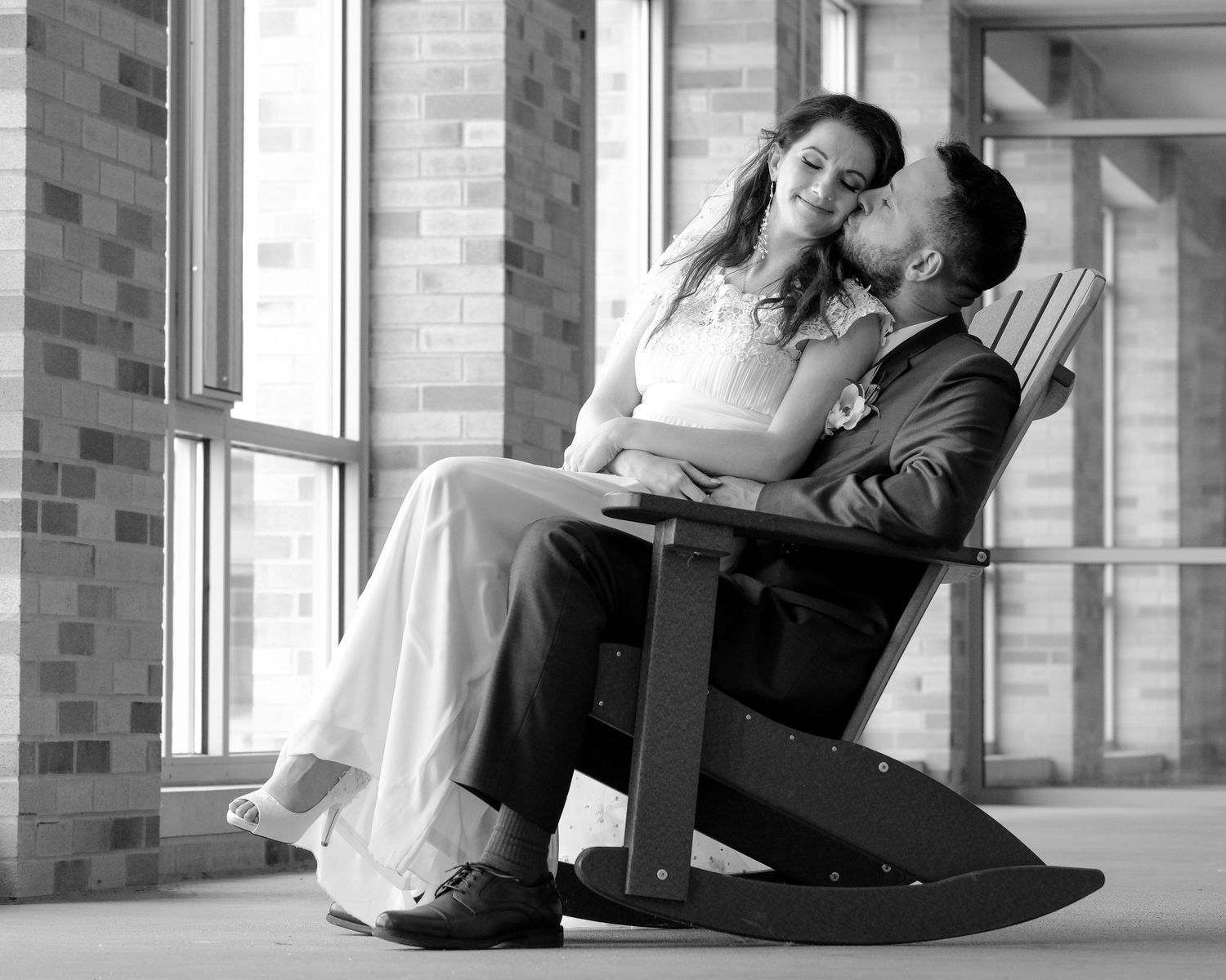 Black and white photo of a bride in a flowing gown sitting on a groom's lap in a rocking chair as he kisses her cheek on a porch.