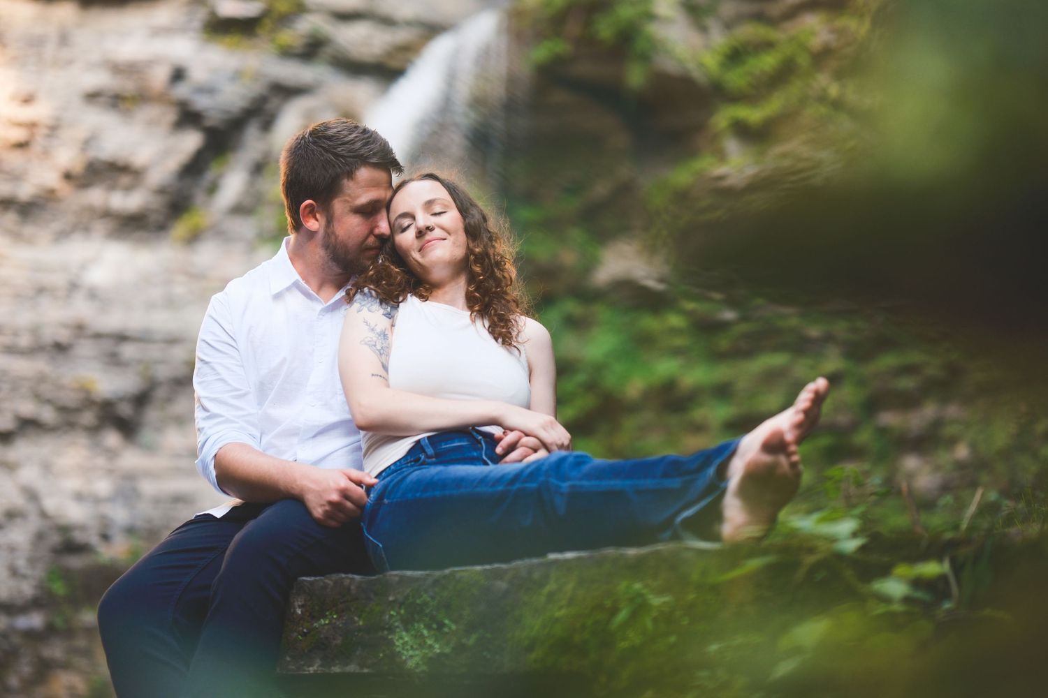 Smiling couple cuddling on a mossy rock ledge in front of a waterfall during a romantic outdoor engagement photo session.