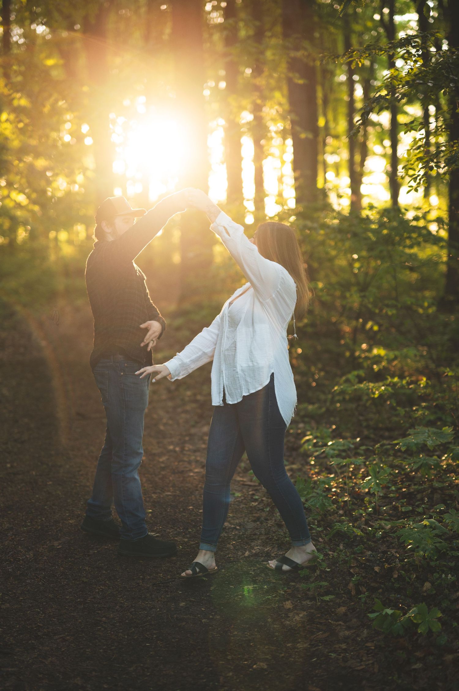 Couple dancing hand in hand on a forest path as golden sunset light streams through the trees.