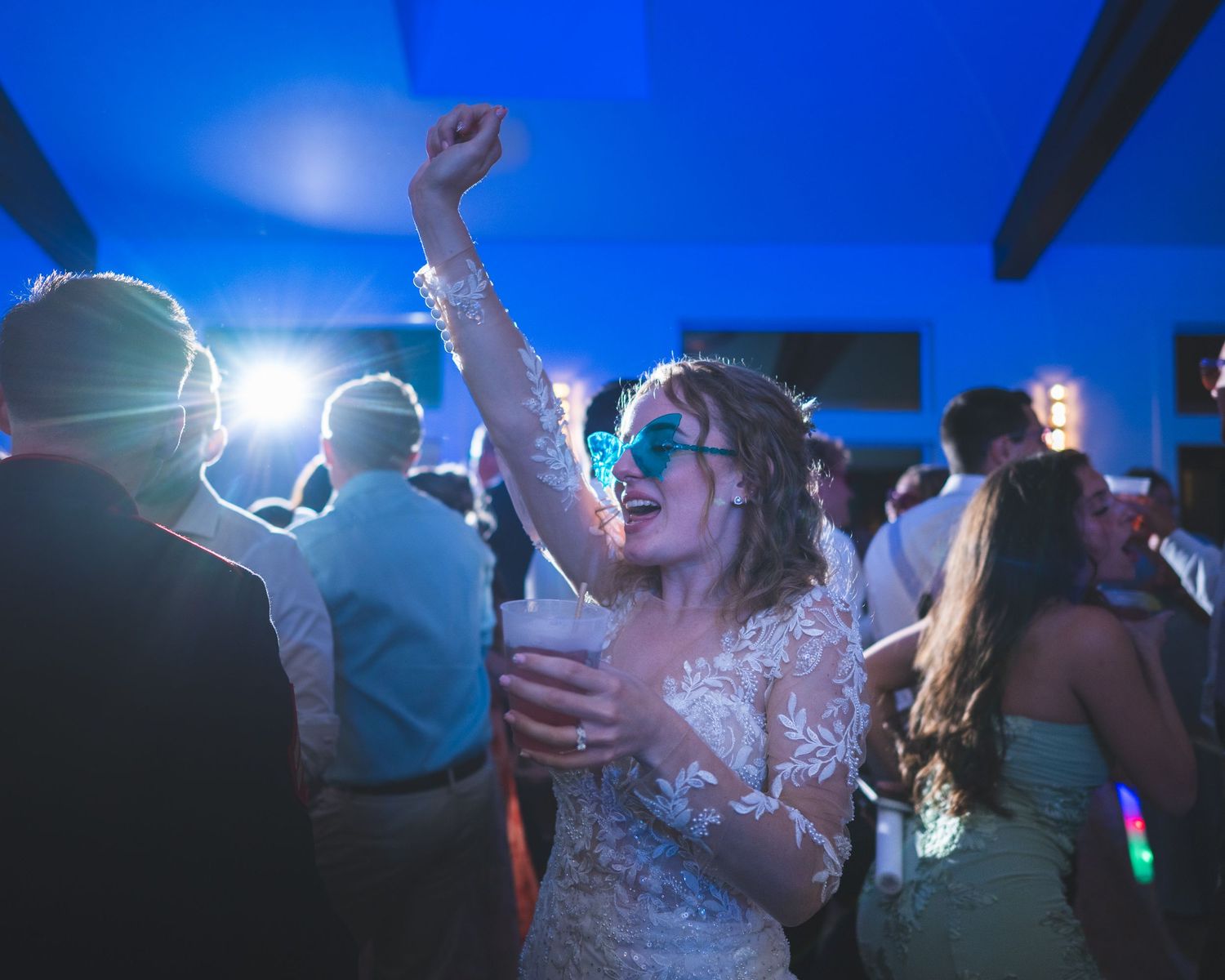 Woman in a white lace dress and blue sunglasses raises her arm and holds a cup while dancing at a lively party with guests behind her.