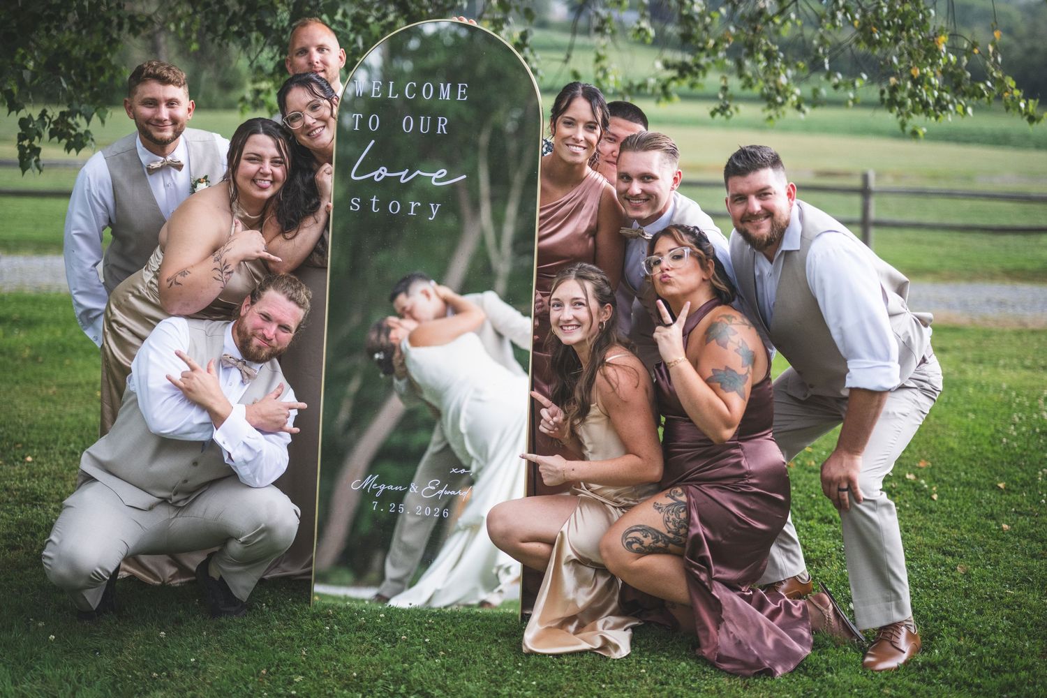 Wedding party laughs and poses around a mirror sign reading welcome to our love story, with bride and groom in the center.