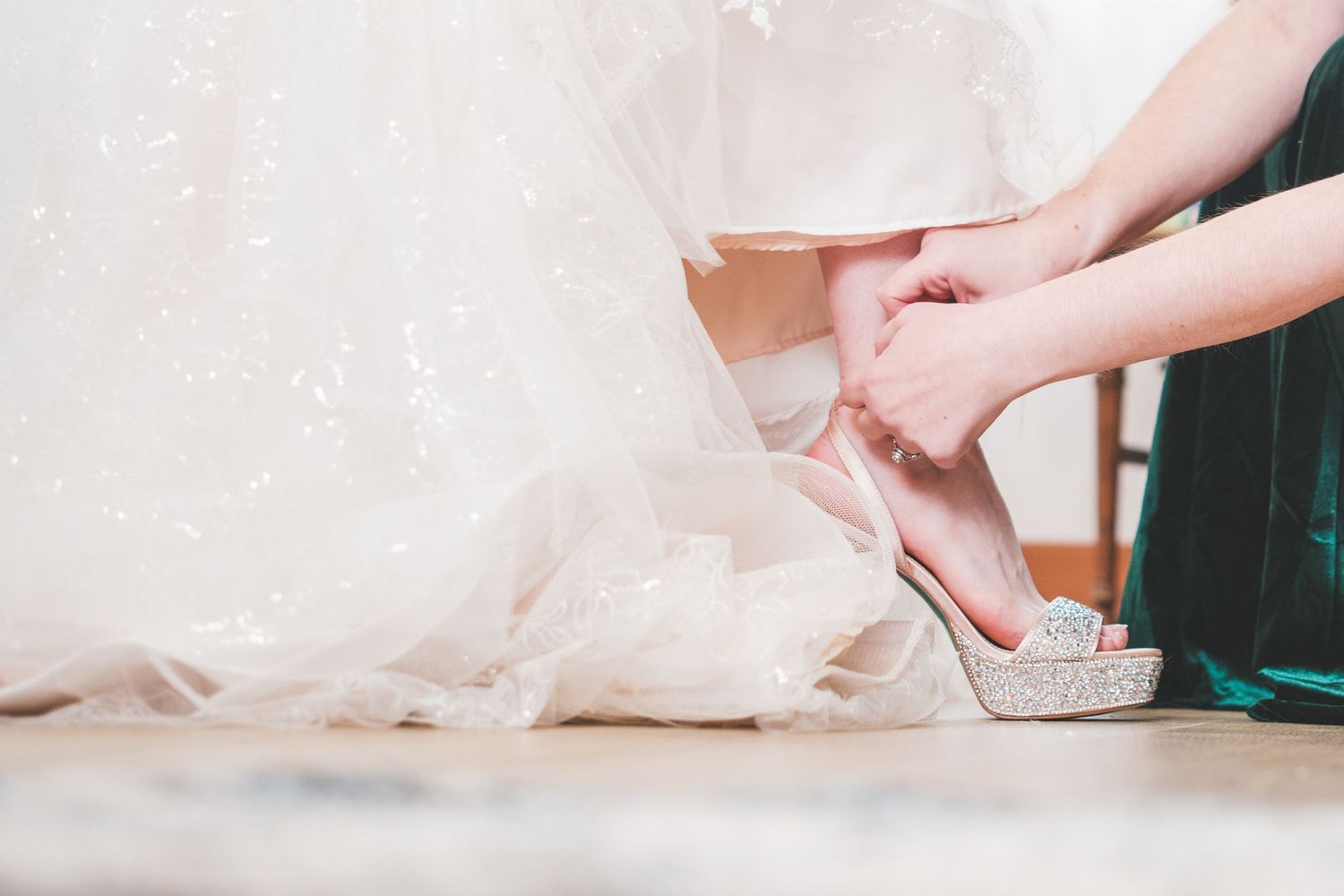 Bridesmaid in green dress helps a bride in a flowing white tulle gown put on a sparkly silver glitter high-heeled wedding shoe.