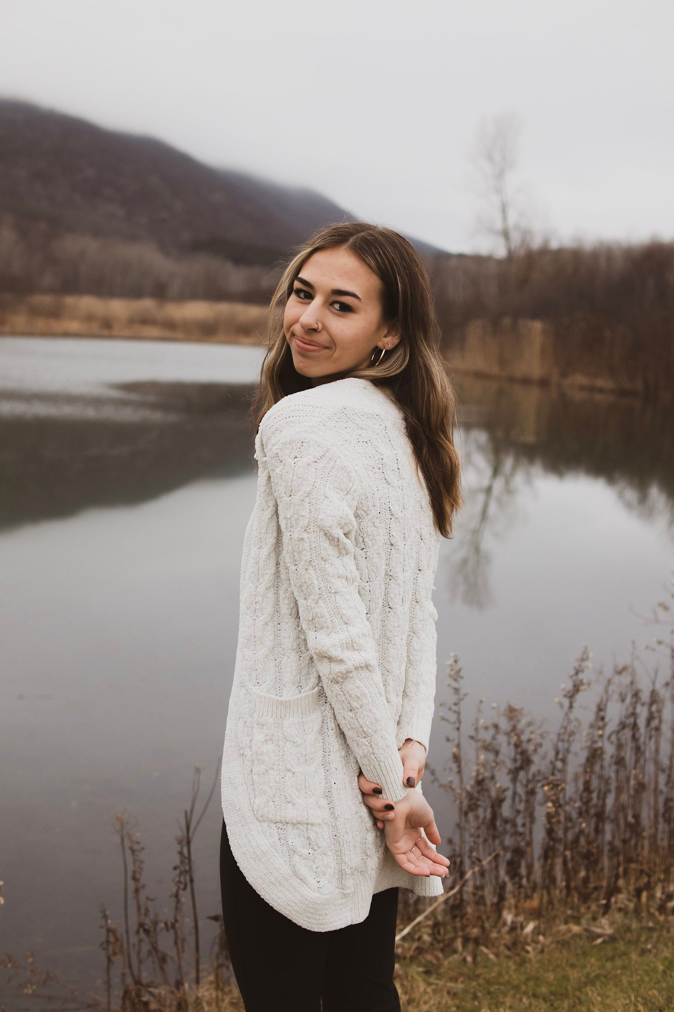 A person wearing a cozy white cable knit sweater poses by a serene lake on an overcast autumn day.
