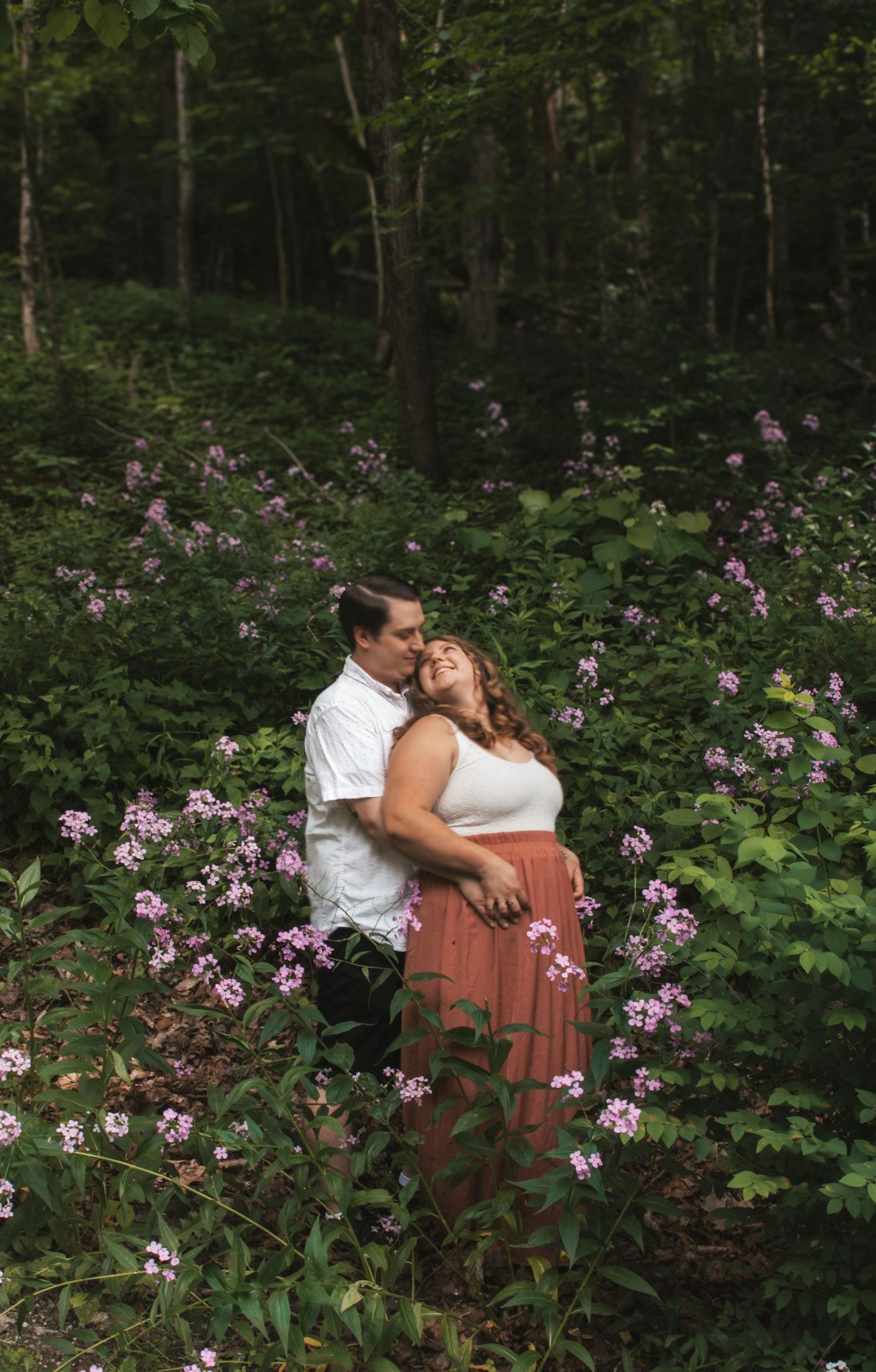 A couple shares a romantic moment while sitting on a wooden bench surrounded by blooming wildflowers in a forest setting.